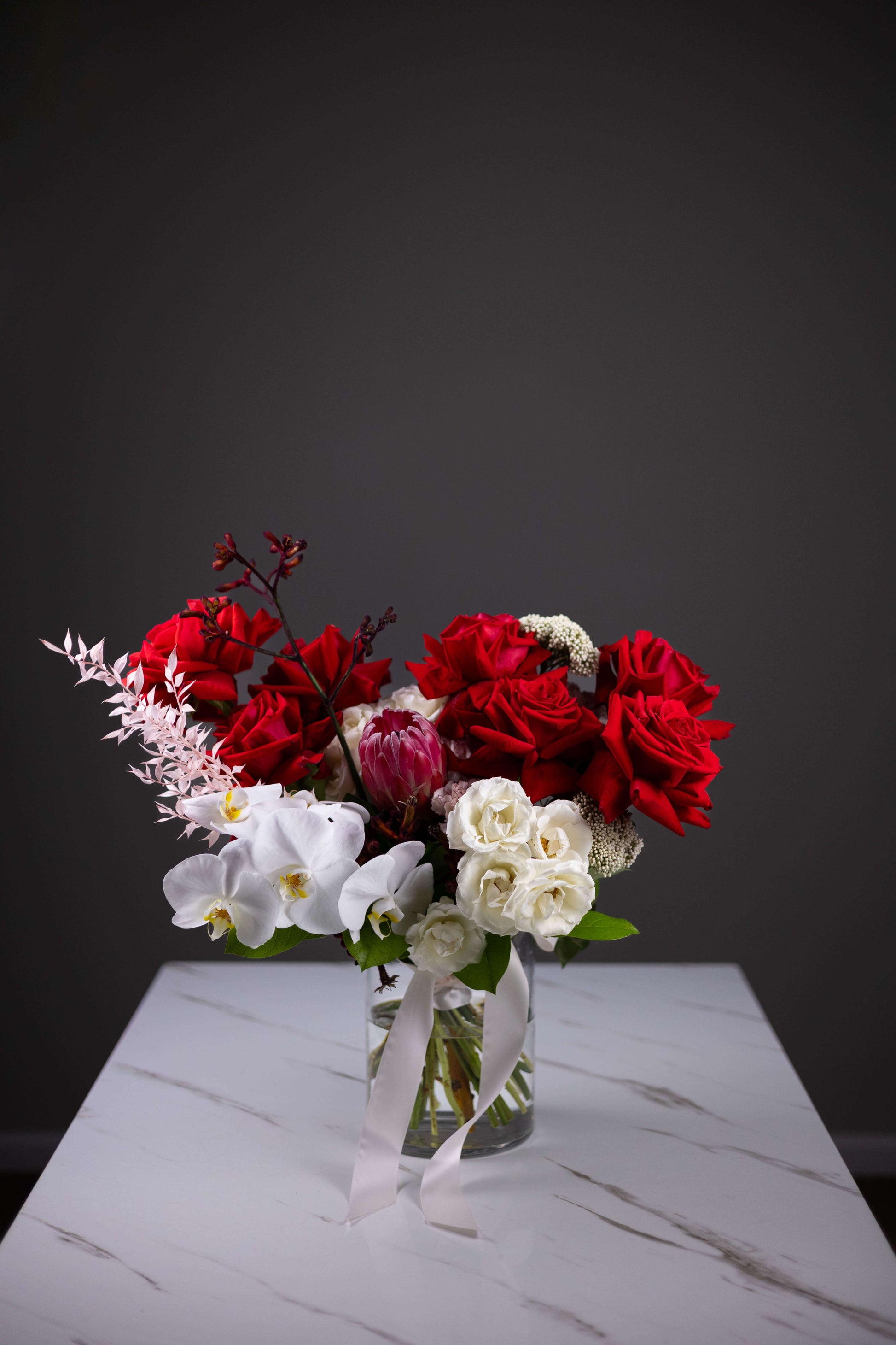 Bouquet of red, white, and pink flowers in a clear vase on a marble surface with a dark background