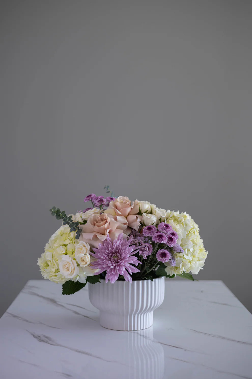 A soft pastel arrangement with cream and blush roses, yellow spray roses, hydrangeas, lavender chrysanthemums, and purple asters. Gentle, fresh, and perfect for any warm occasion.