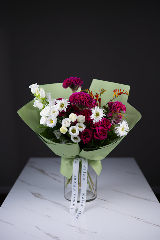 A vibrant hand-tied bouquet with magenta cockscomb, white lisianthus, fuchsia roses, and fiery crocosmia, wrapped in light green paper and placed in a clear glass vase.