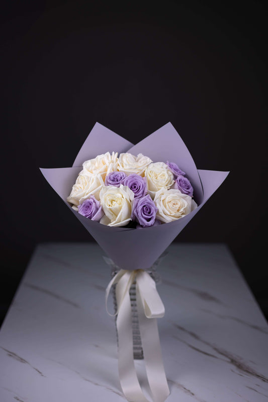 Bouquet of white and purple roses wrapped in purple paper with a white ribbon on a dark background.