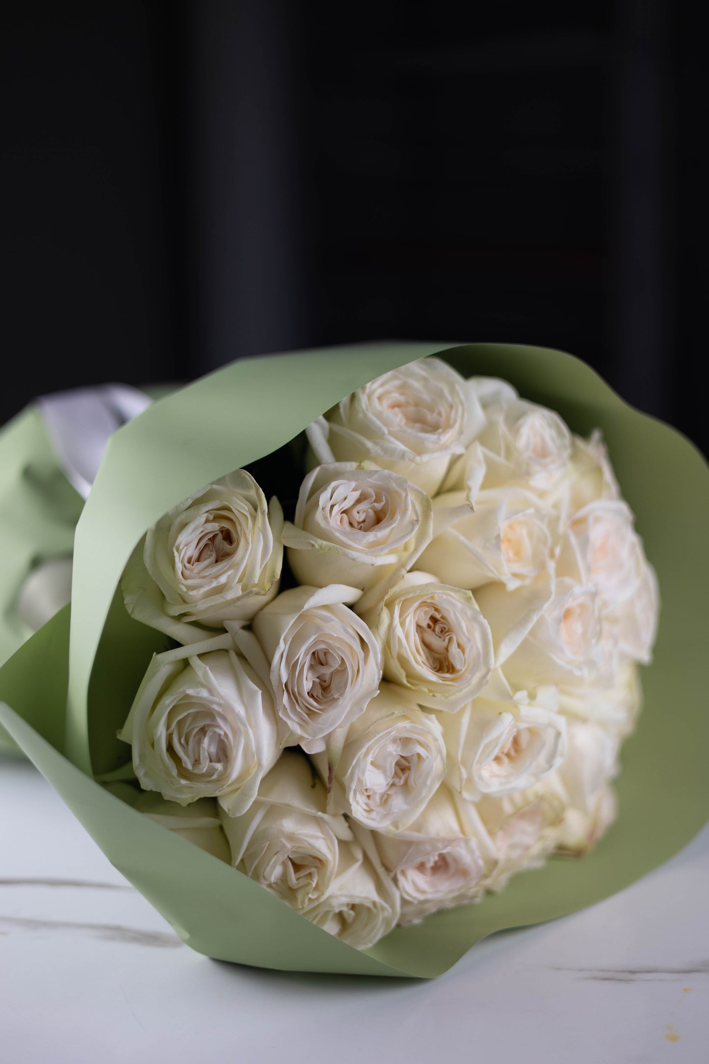 Close-up view of a hand-tied bouquet of white roses with a soft blush center, wrapped in muted sage-green paper.