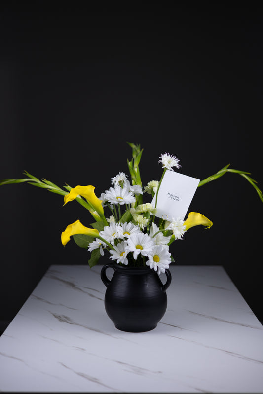 Full view of a bright spring arrangement with daisies and yellow calla lilies in a black vase on a white marble table, dark background.