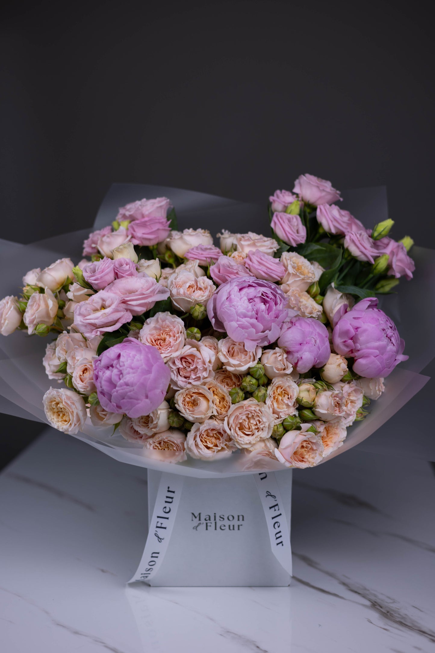 Delicate bouquet of blush spray roses, lavender lisianthus, and soft pink peonies wrapped in translucent paper, standing in a Maison d’Fleur branded glass vase.
