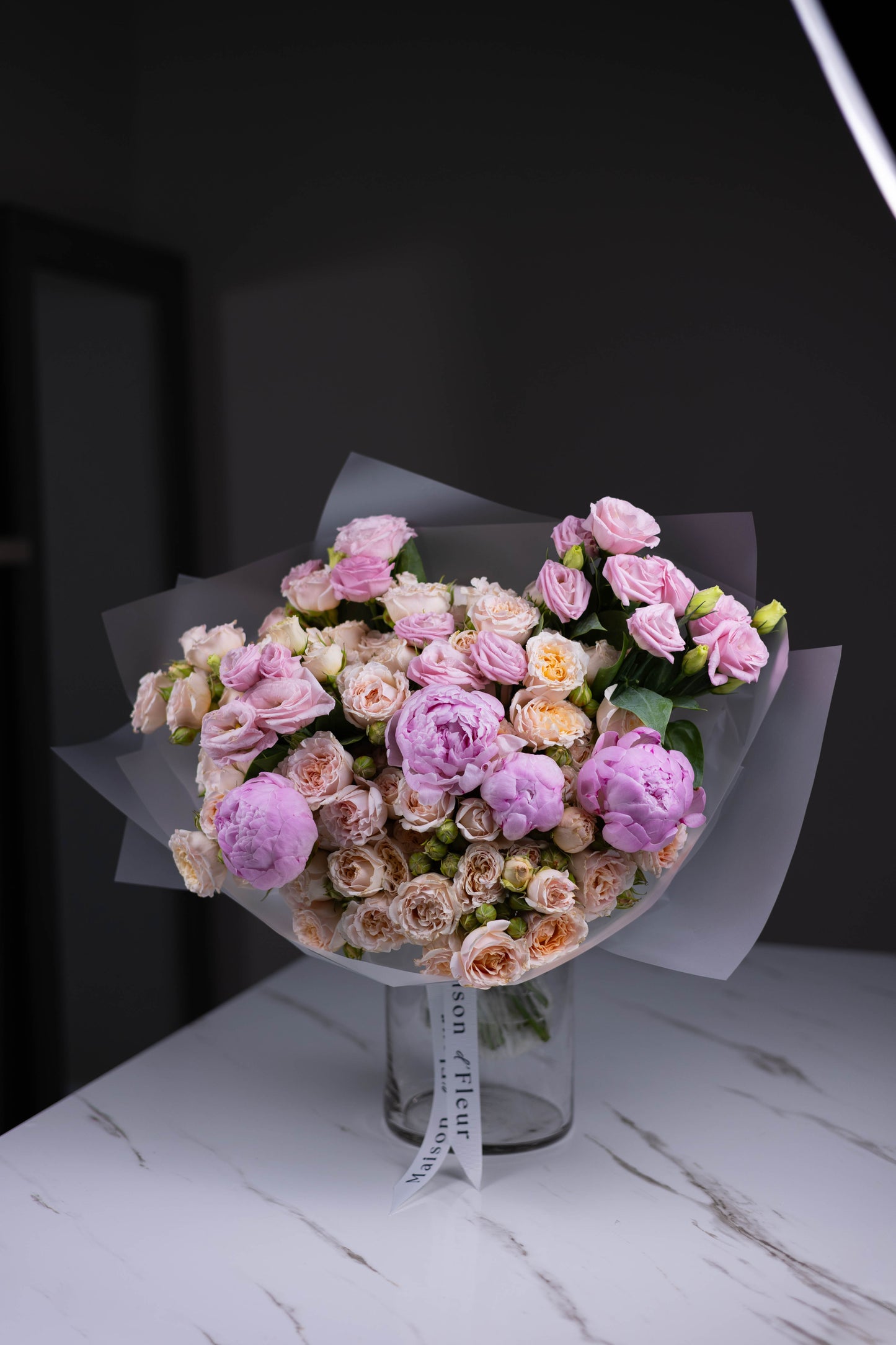 Pastel flower arrangement with pink peonies, cream spray roses, and lavender lisianthus displayed in a clear vase with branded ribbon.