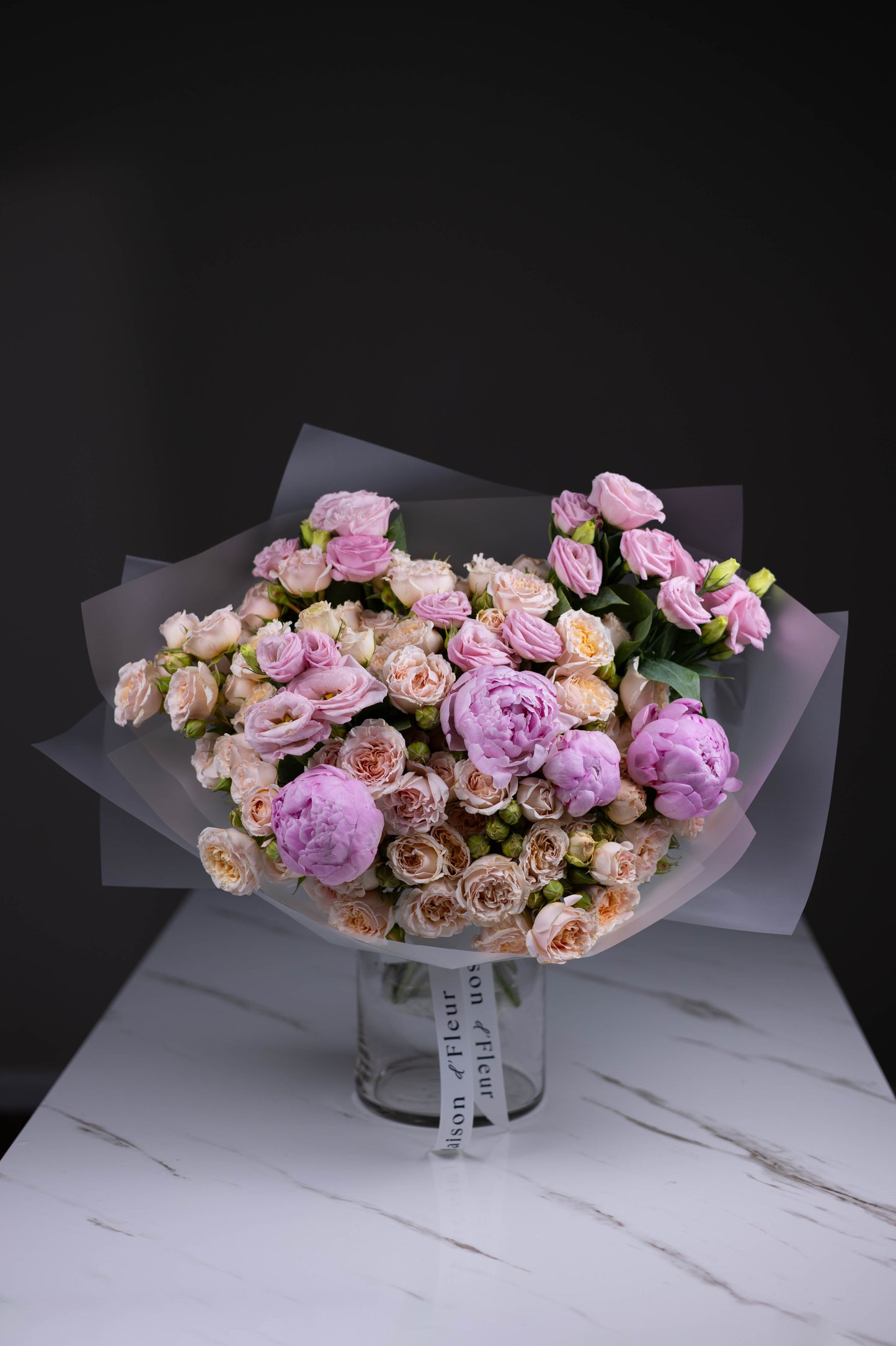 Delicate bouquet of blush spray roses, lavender lisianthus, and soft pink peonies wrapped in translucent paper, standing in a Maison d’Fleur branded glass vase.