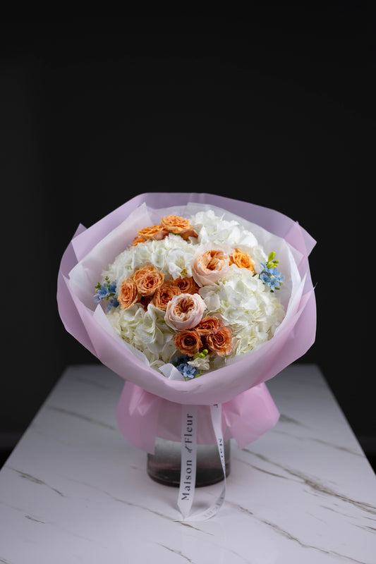 Delicate bouquet with white and peach-toned flowers wrapped in pastel pink paper, placed on a marble surface under studio lighting, featuring Maison d’Fleur branded ribbon.