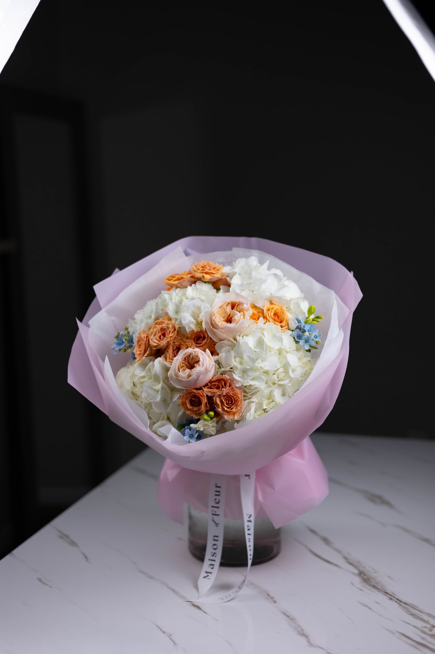 Front view of a voluminous bouquet with creamy white hydrangeas, soft pink garden roses, and pops of sky blue and lime green blooms, styled in pink paper wrapping.