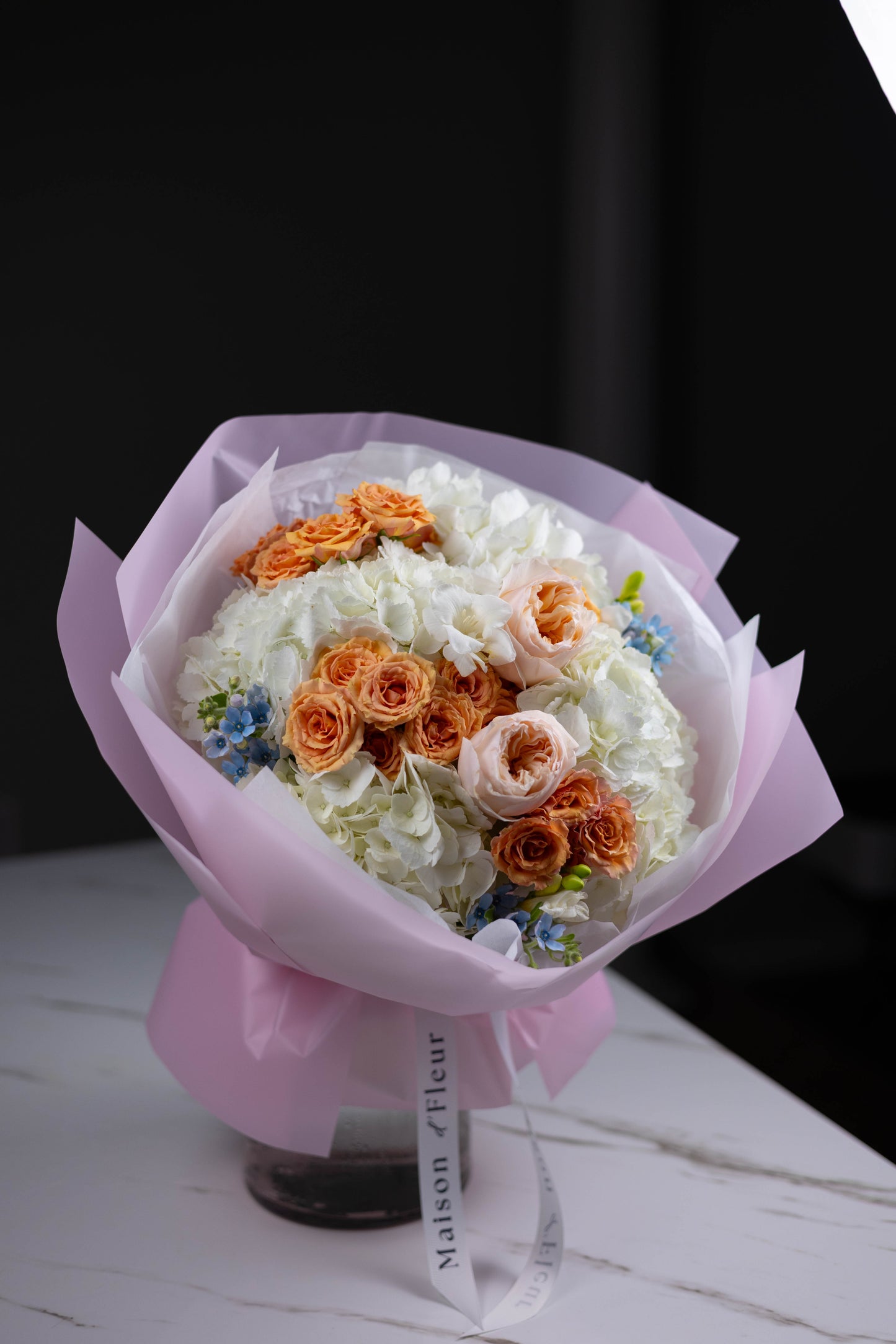 Angled shot of a round bouquet with white hydrangeas and a mix of peach, blush, and orange spray roses, complemented by pastel blue floral details.