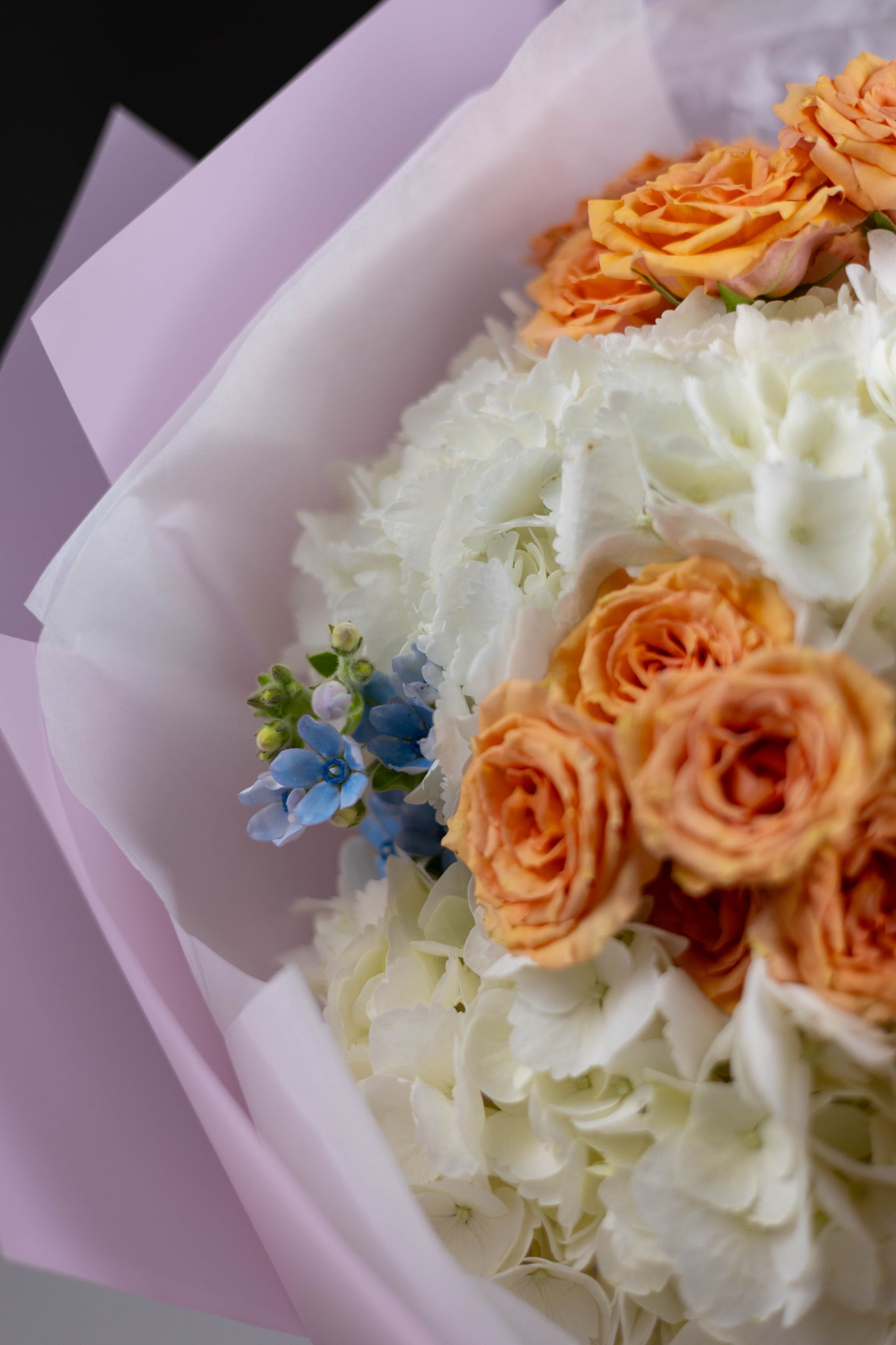 Side view of a romantic bouquet featuring fluffy white hydrangeas and vivid orange-peach roses, accented with clusters of sky-blue flowers.