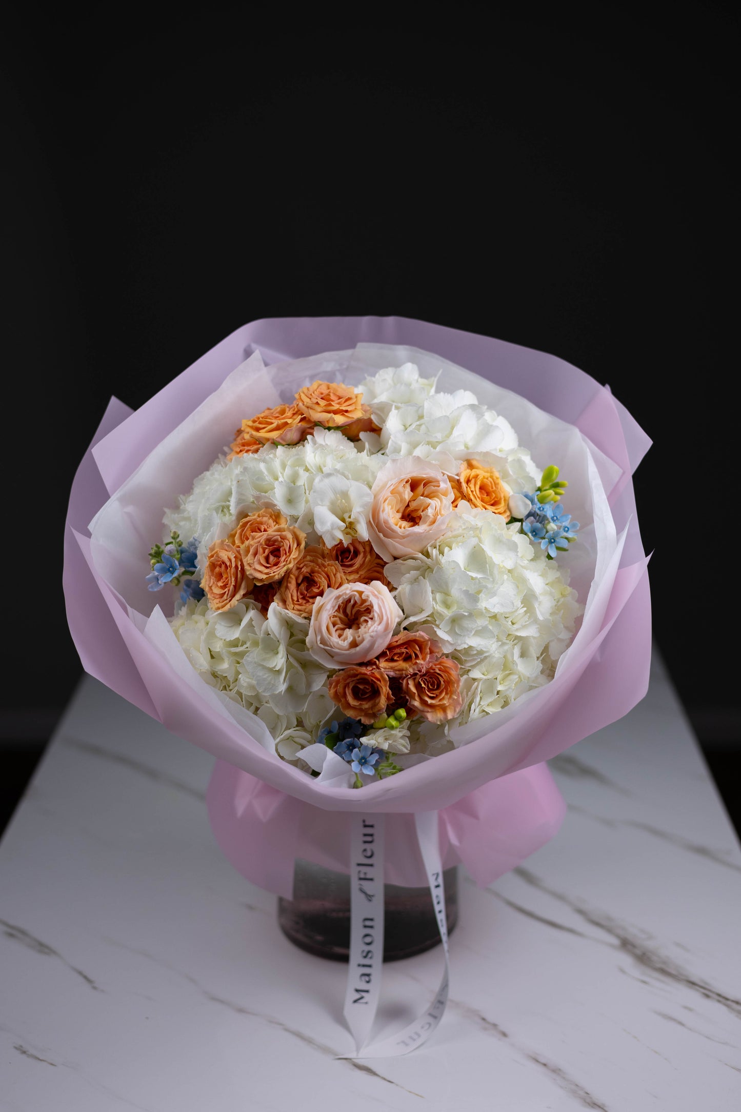 Full bouquet of white hydrangeas, peach and blush roses, and delicate blue accents, wrapped in light pink paper with Maison d’Fleur ribbons, placed on a marble table.