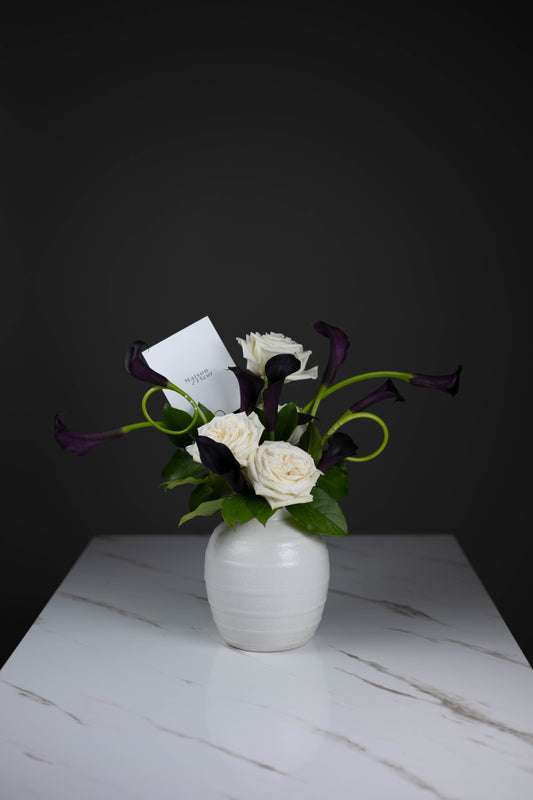 Designer floral piece in white ceramic vase featuring white garden roses and black calla lilies