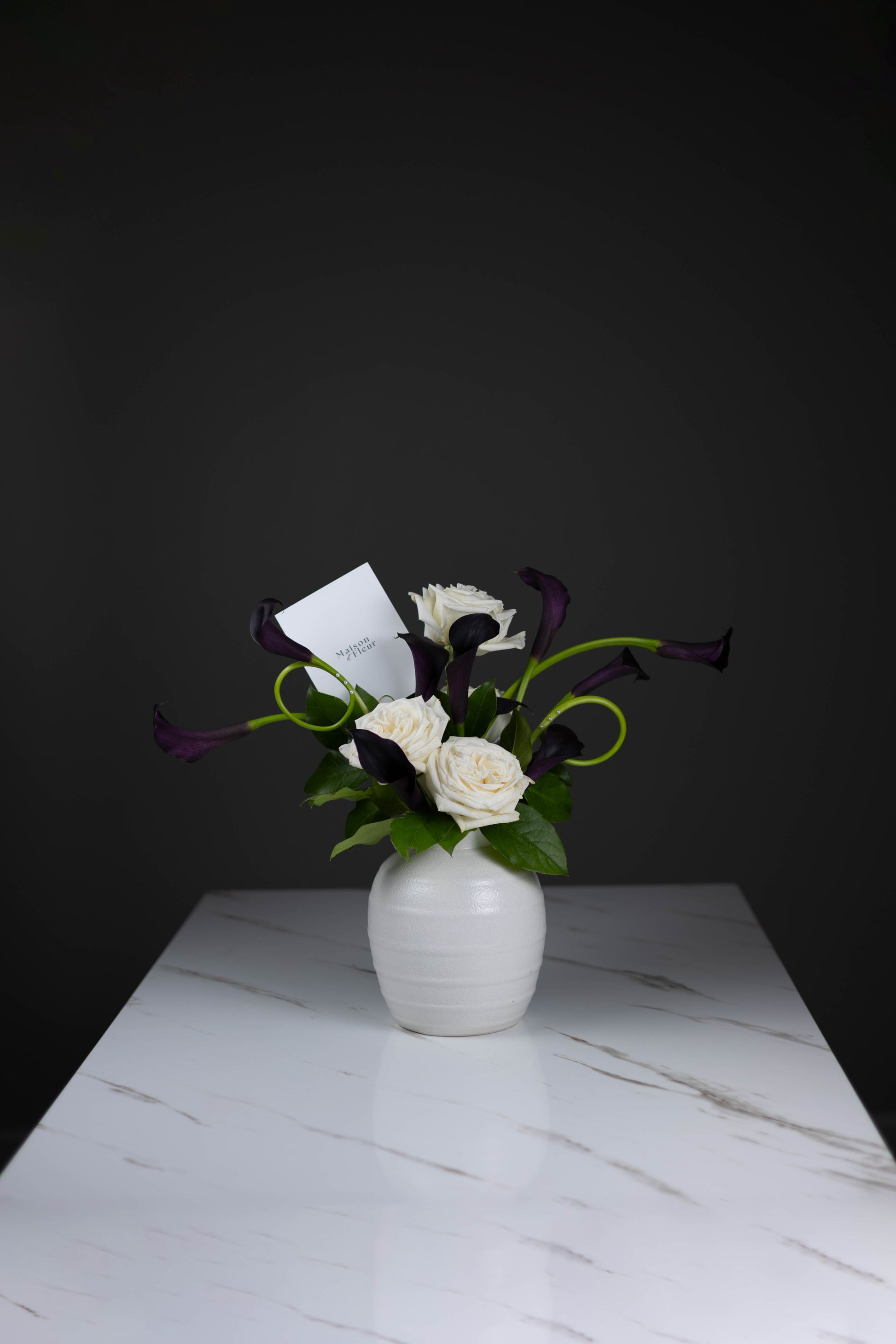 High-end floral arrangement in white vase with cream roses and dark calla lilies on marble table