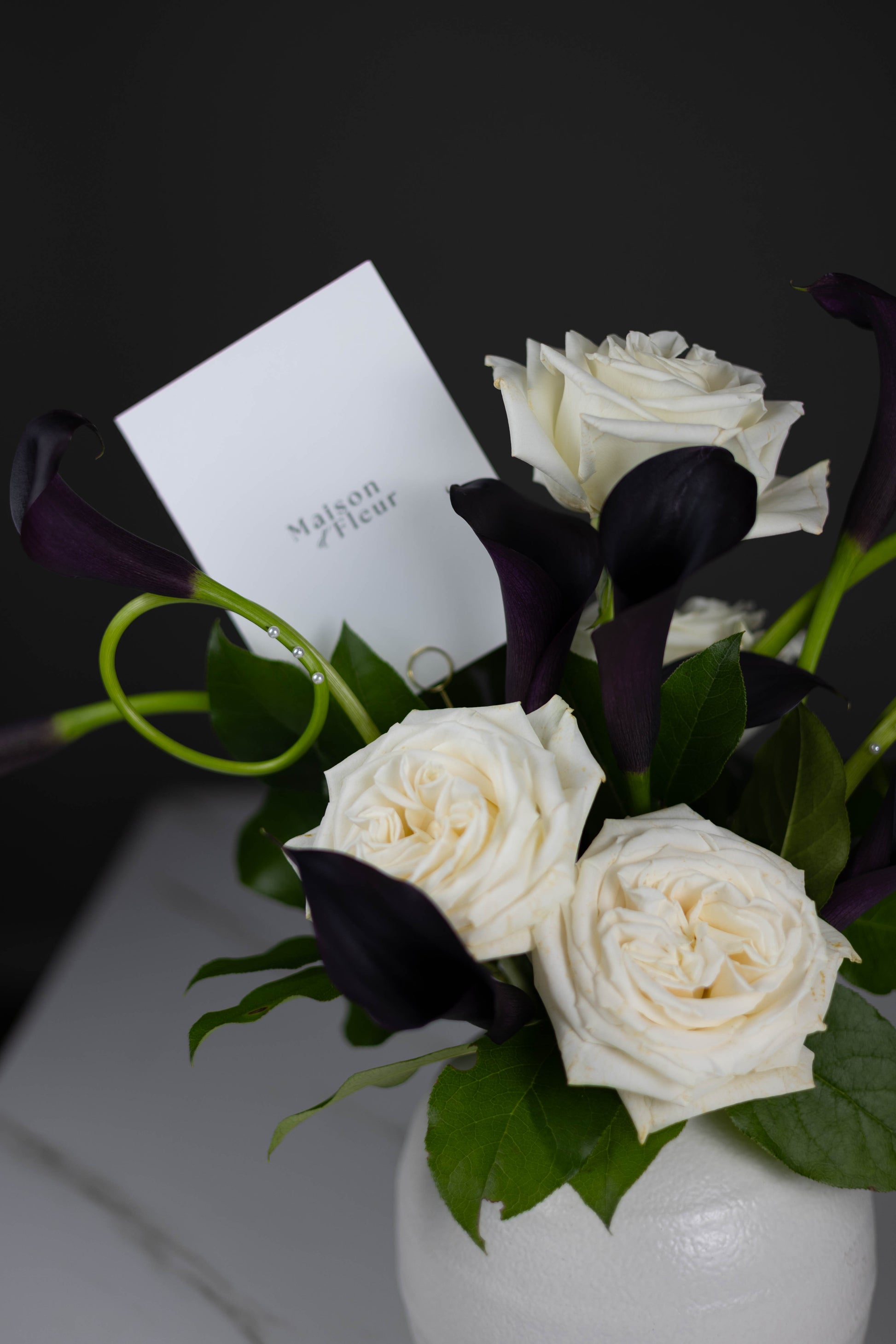 White roses and deep plum calla lilies arranged with Maison d’Fleur card in black background