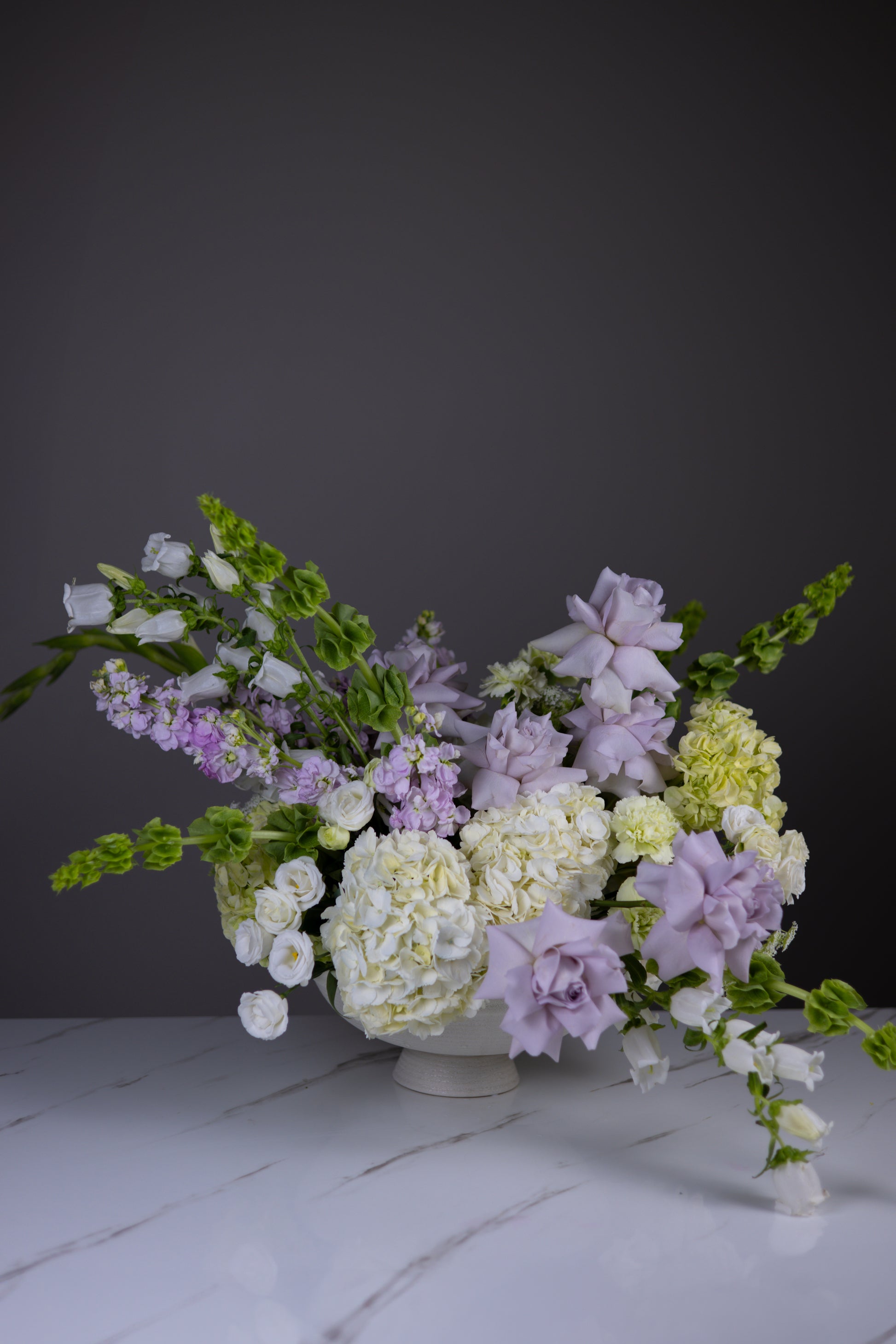A luxurious garden-style centerpiece with lavender roses, white hydrangeas, and cascading greenery in a ceramic compote vase.