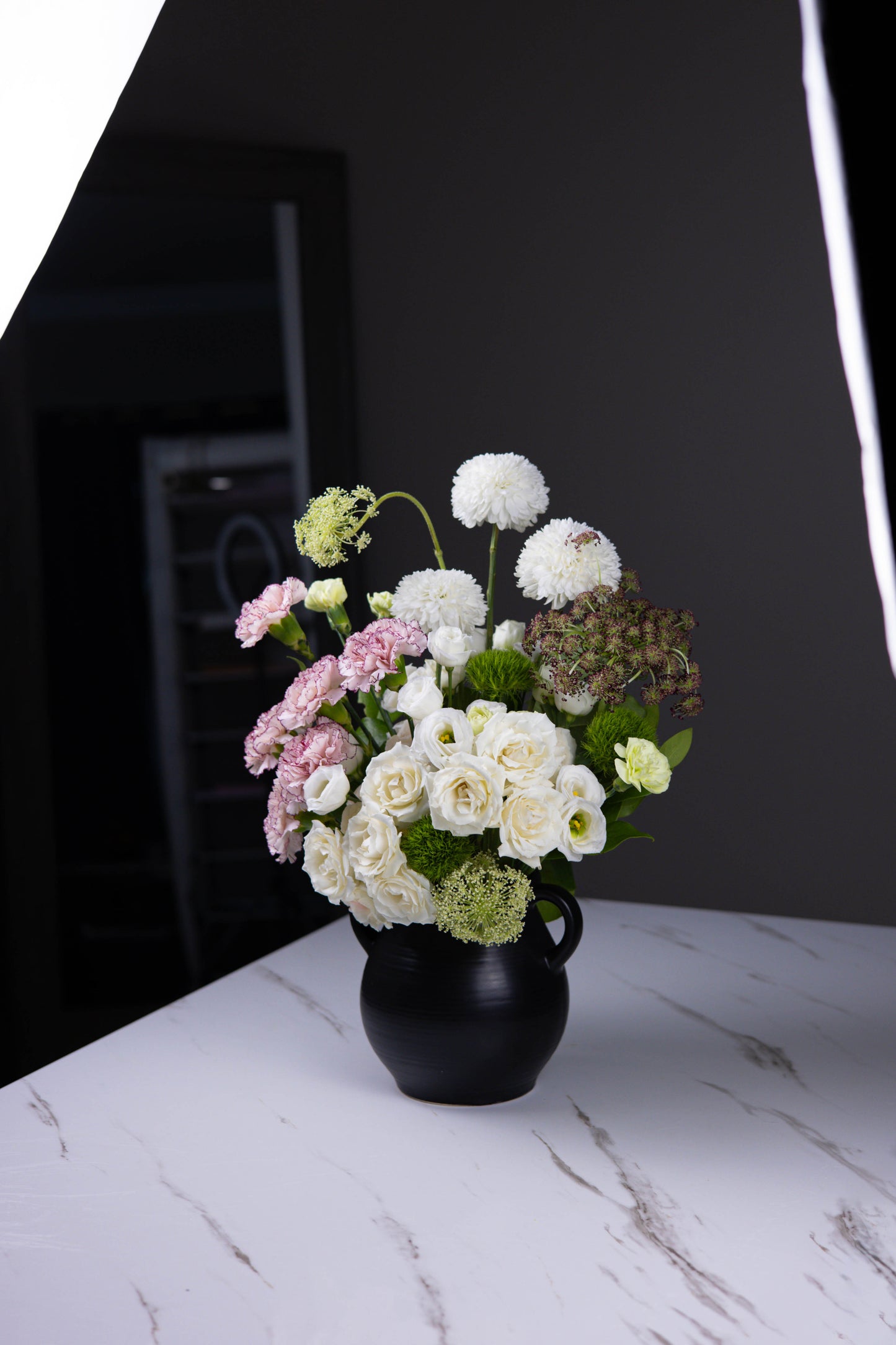 Ivory and blush floral arrangement with white roses, carnations, and dahlias in a black ceramic vase on a marble table