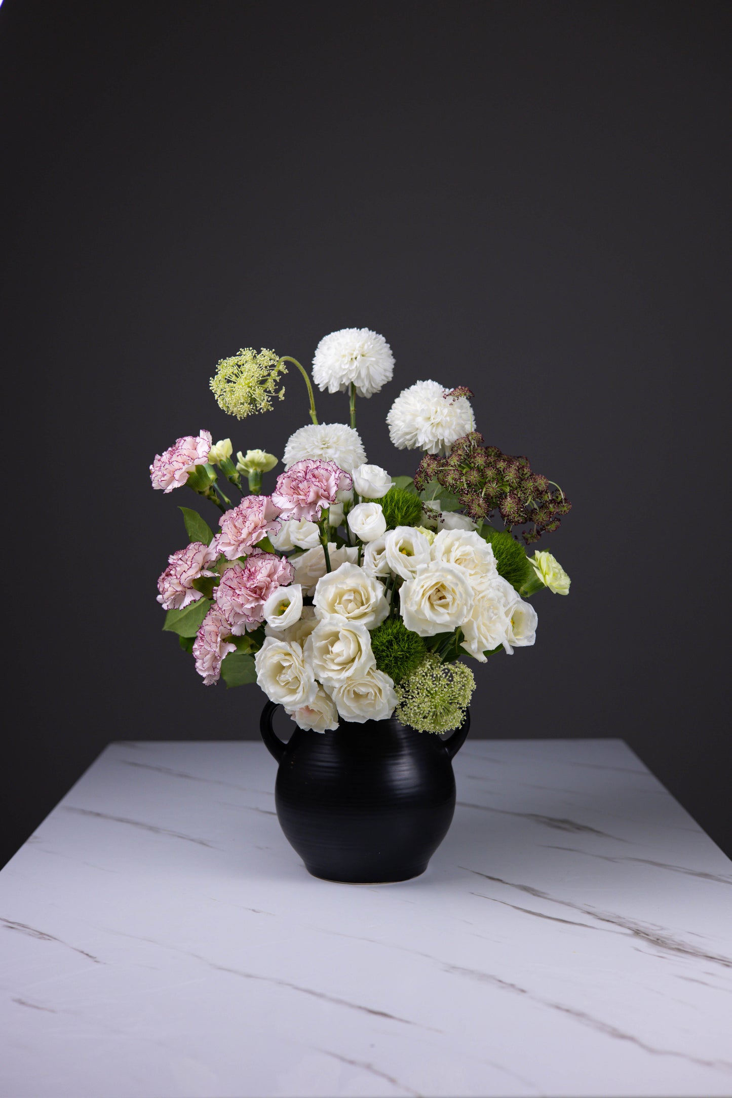 Front-facing ivory bouquet with roses, pink carnations, and green textures in a black jug vase