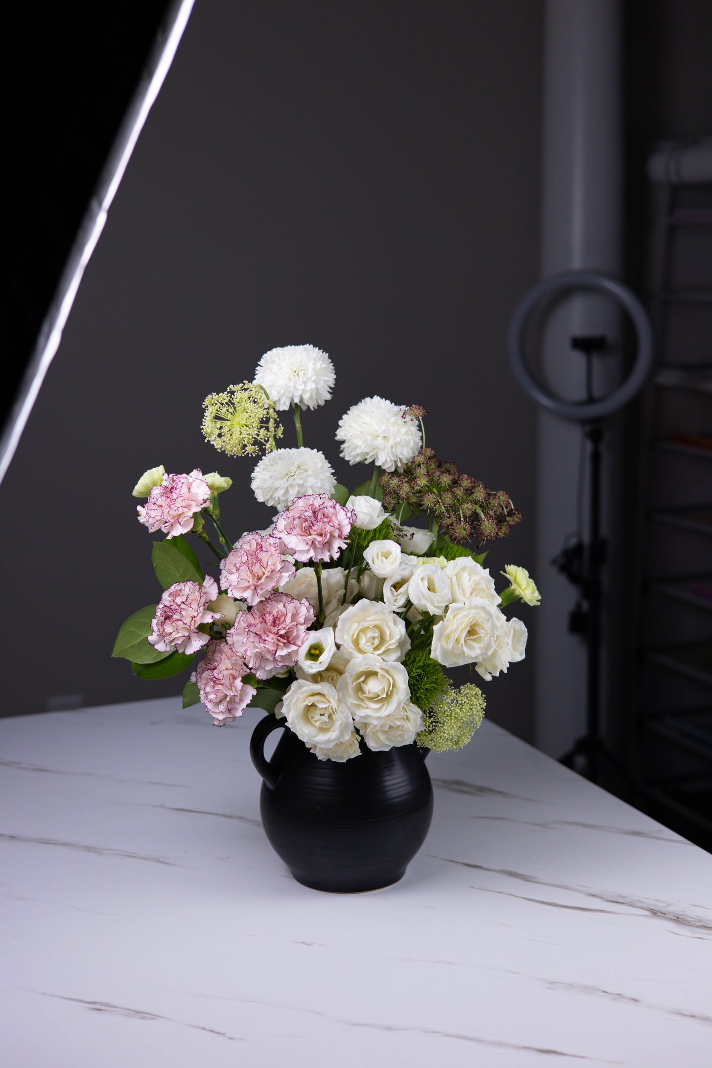 Side view of a white and pink floral bouquet styled in a modern black vase against a dark gray backdrop