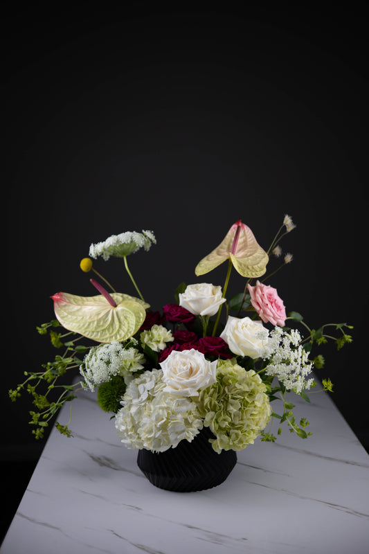 Full front view of modern garden-style arrangement in a black vase, showcasing roses, hydrangeas, and anthuriums.