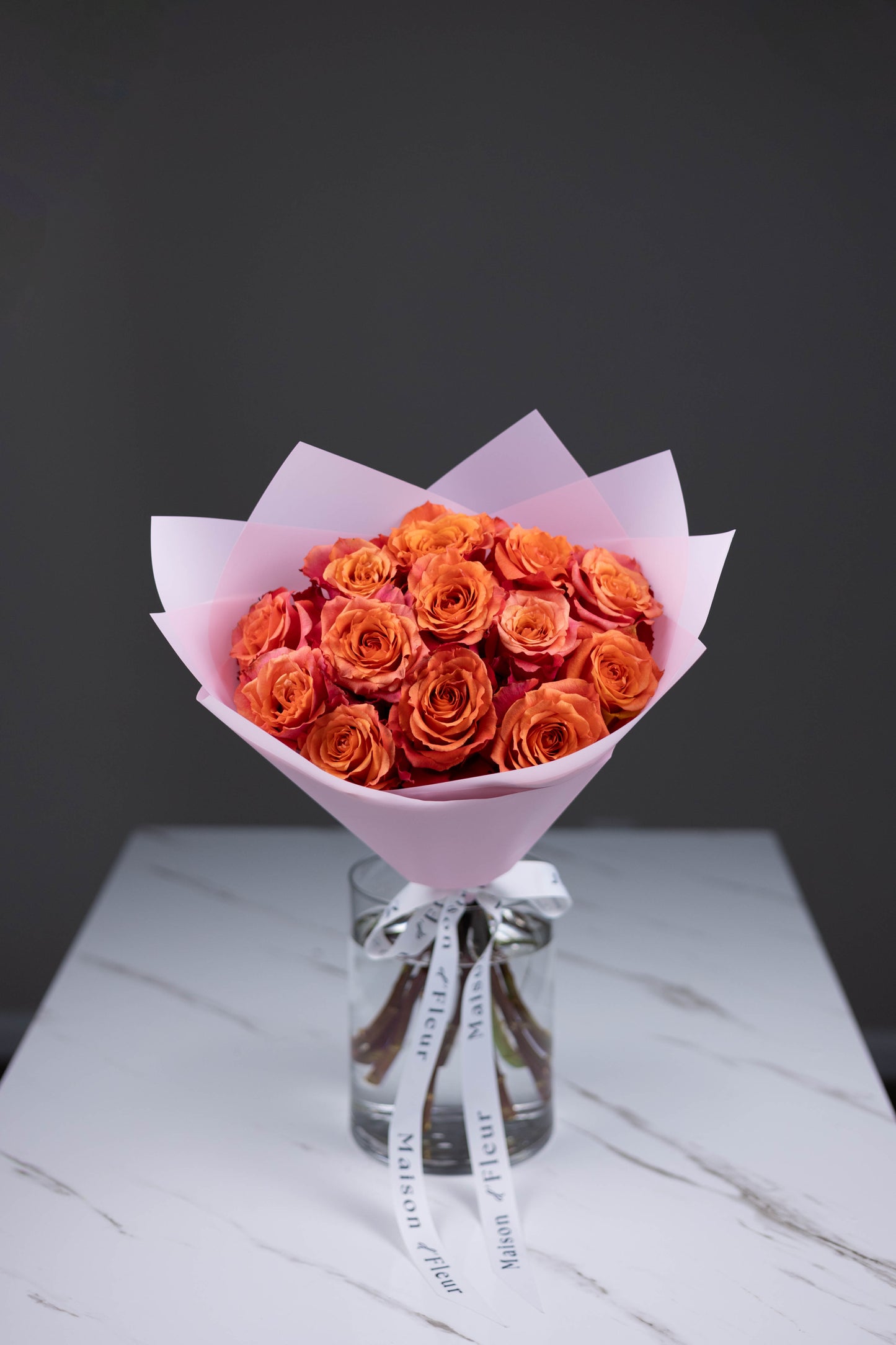 A hand-tied bouquet of vibrant orange-pink Nature’s Cherry roses, arranged in soft pink wrapping paper with Maison d’Fleur custom ribbon, placed in a clear glass vase on a white marble surface.