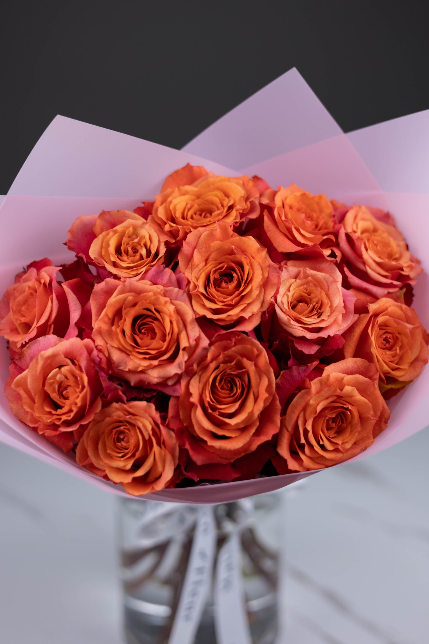 A hand-tied bouquet of vibrant orange-pink Nature’s Cherry roses, arranged in soft pink wrapping paper with Maison d’Fleur custom ribbon, placed in a clear glass vase on a white marble surface.