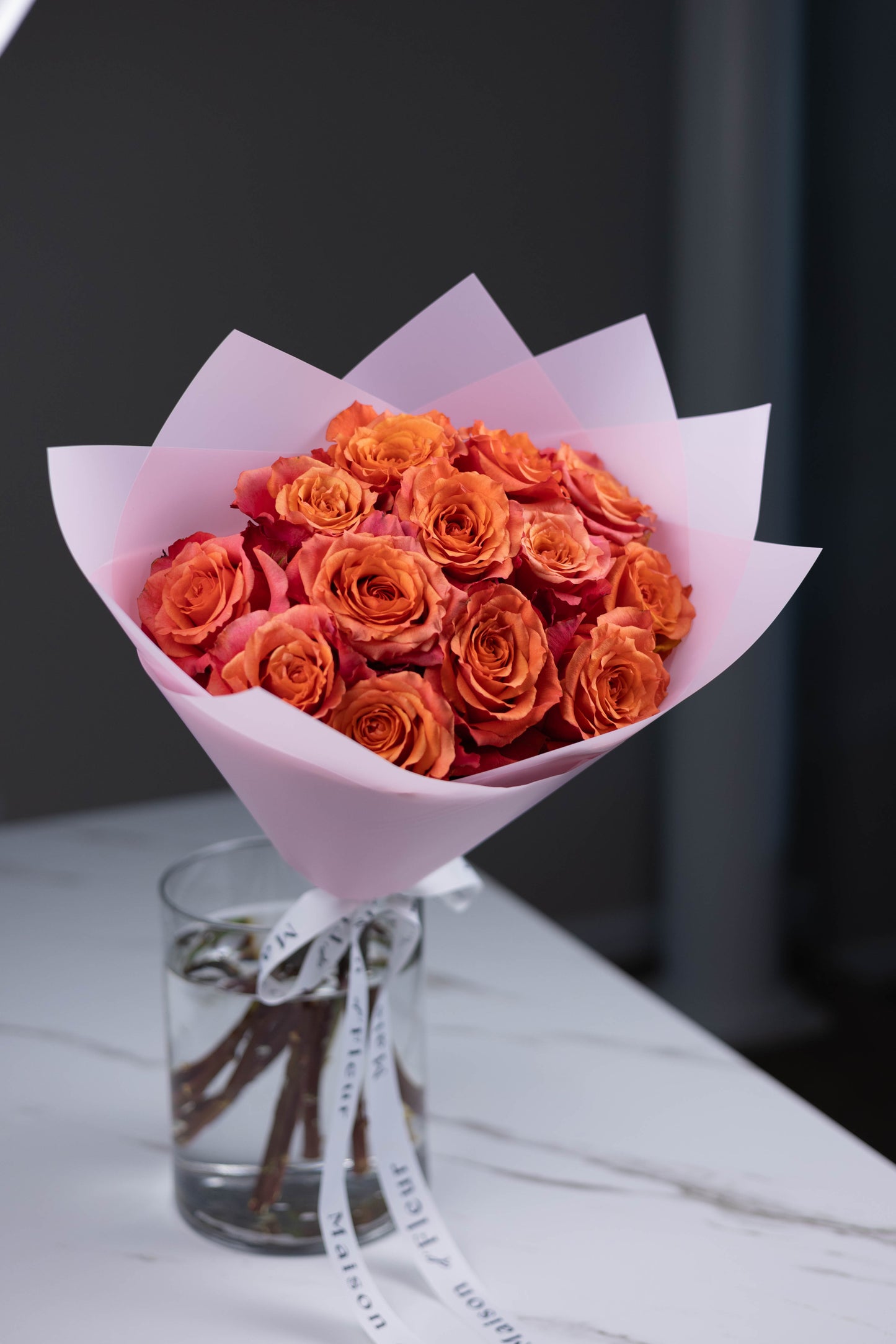A hand-tied bouquet of vibrant orange-pink Nature’s Cherry roses, arranged in soft pink wrapping paper with Maison d’Fleur custom ribbon, placed in a clear glass vase on a white marble surface.