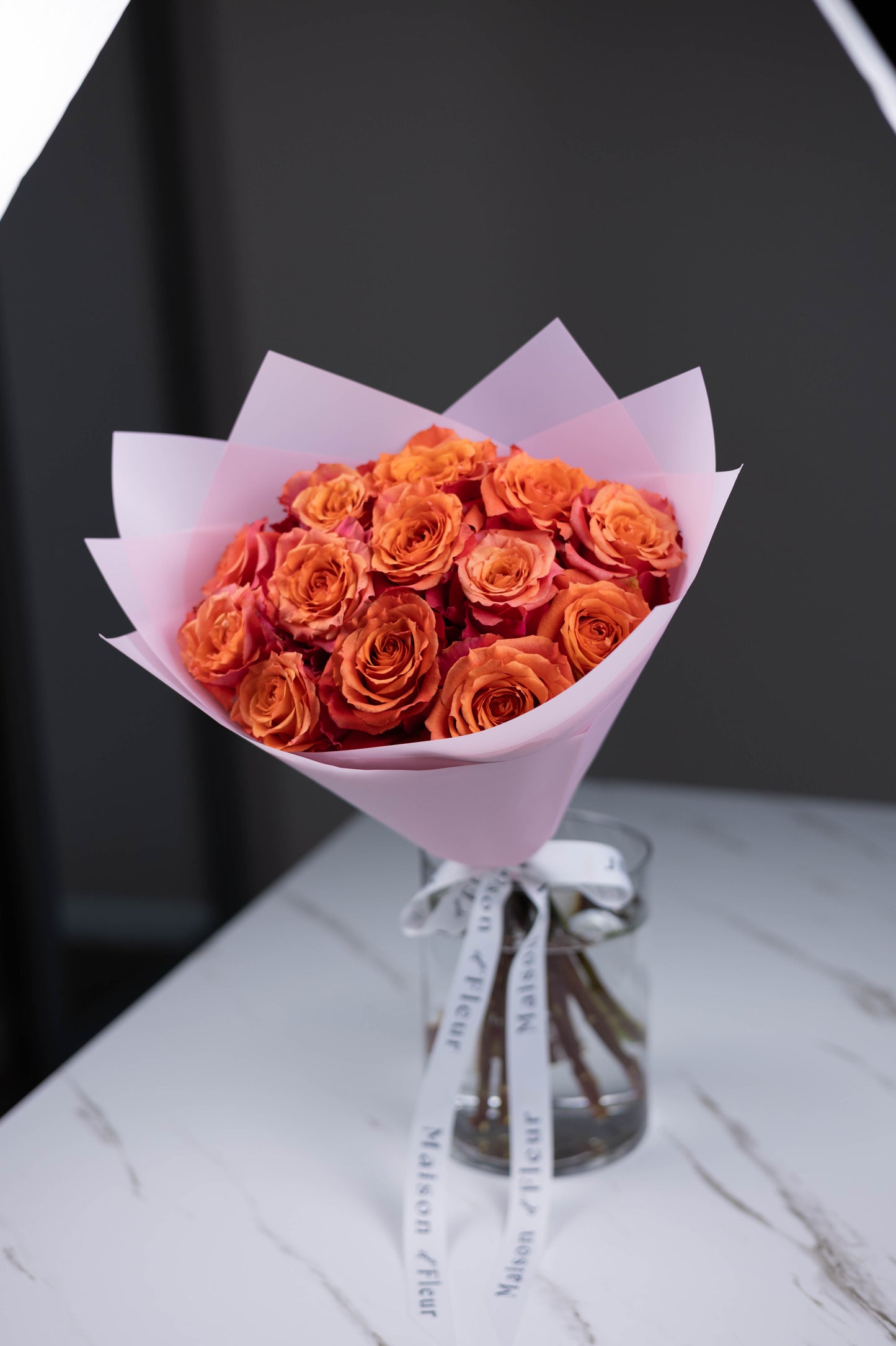 A hand-tied bouquet of vibrant orange-pink Nature’s Cherry roses, arranged in soft pink wrapping paper with Maison d’Fleur custom ribbon, placed in a clear glass vase on a white marble surface.