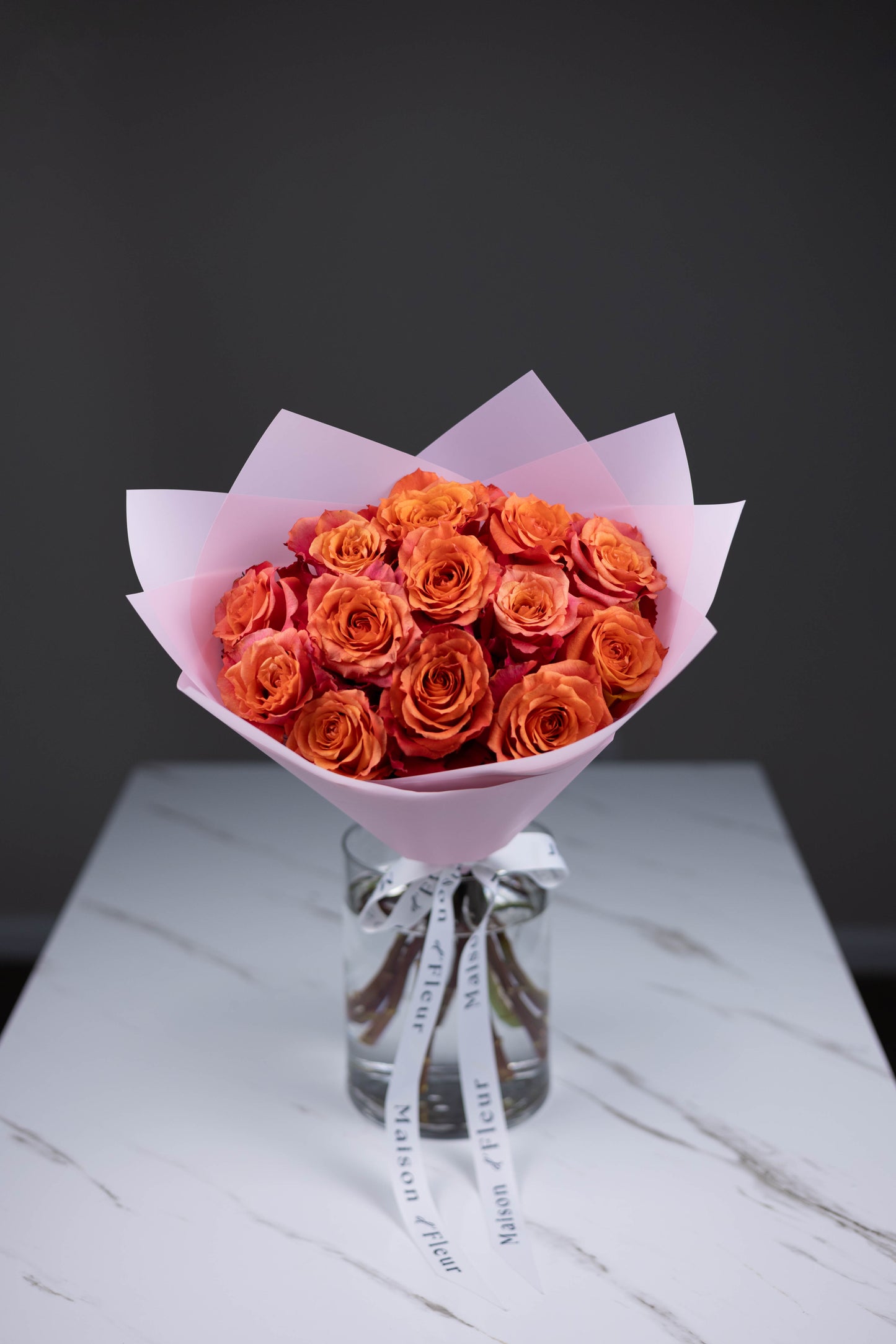 A hand-tied bouquet of vibrant orange-pink Nature’s Cherry roses, arranged in soft pink wrapping paper with Maison d’Fleur custom ribbon, placed in a clear glass vase on a white marble surface.