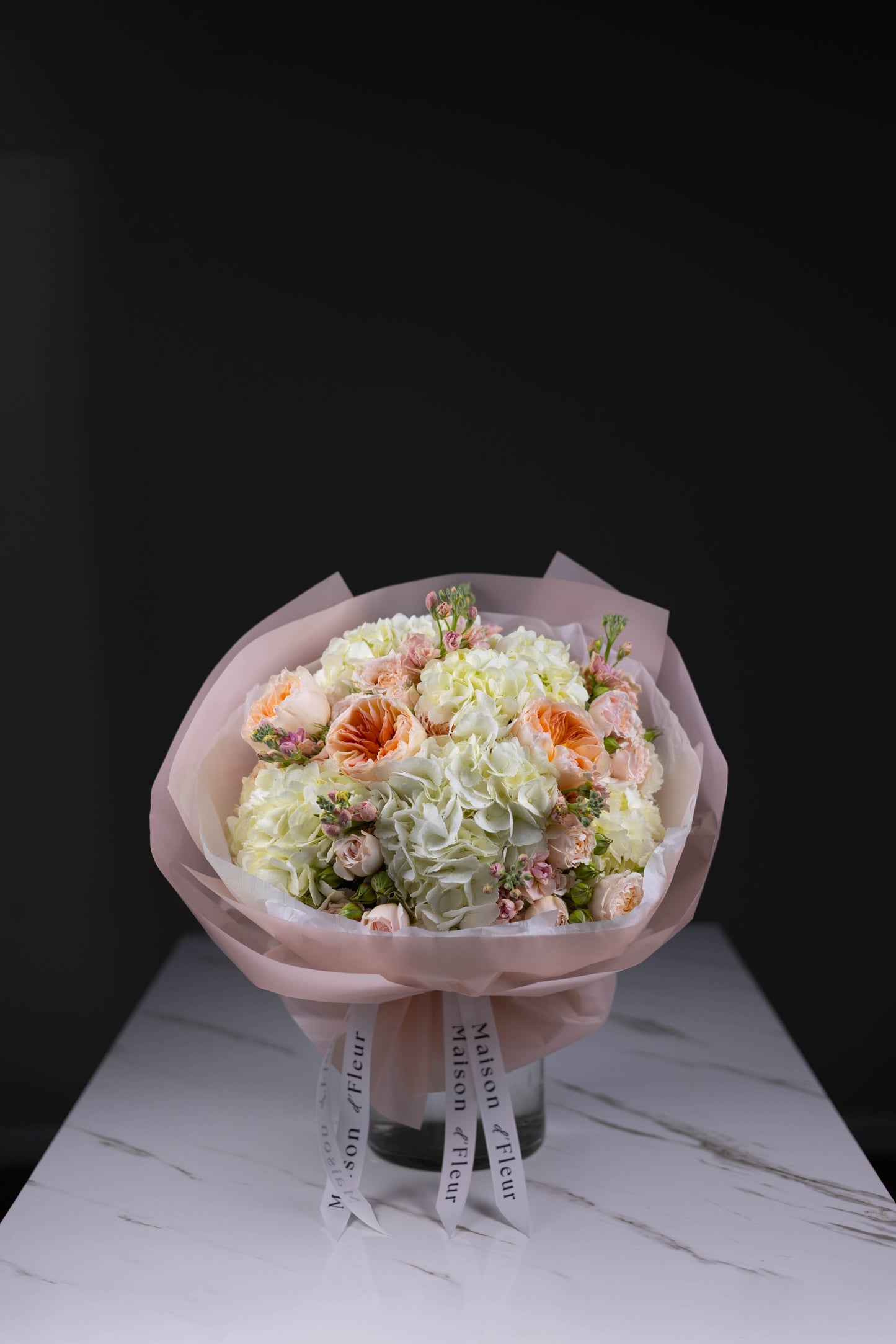 Pastel bouquet featuring white hydrangeas, blush spray roses, stock flowers, and soft peach garden roses, wrapped in pink paper with Maison d’Fleur ribbon, displayed on a white marble table.