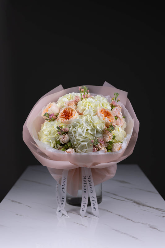 Elegant floral arrangement in blush and cream tones with large white hydrangeas and soft peach accents, presented in pink wrapping and Maison d’Fleur branded ribbons.