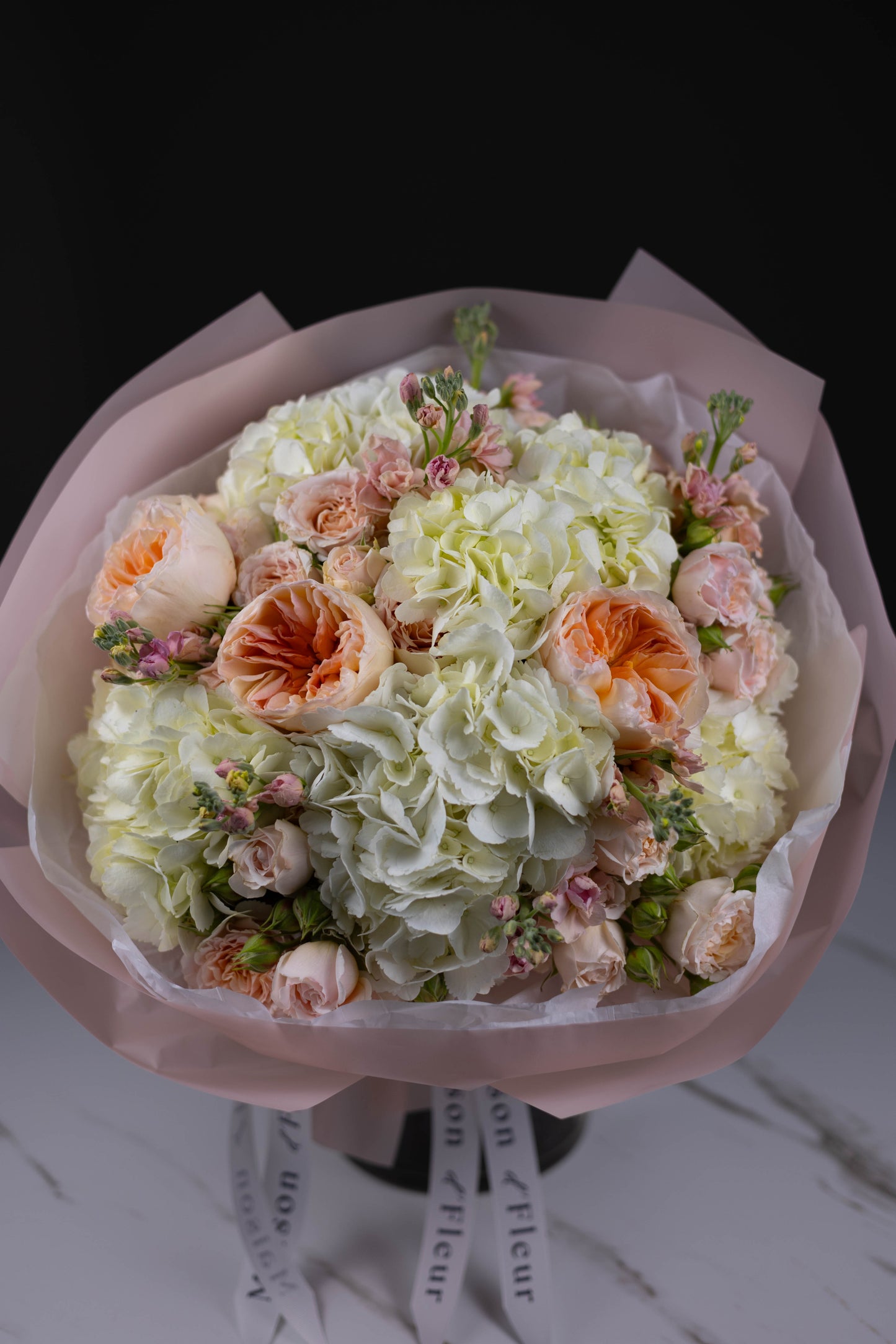Close detail of garden roses and hydrangeas in pastel colors, wrapped in blush-toned paper—elegant and lush, captured against a dark background.