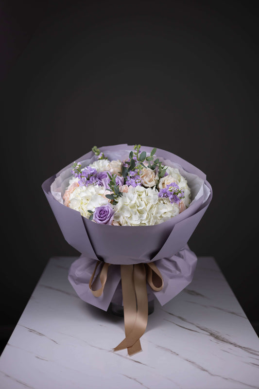 Bouquet of flowers wrapped in purple paper with a brown ribbon on a white surface.
