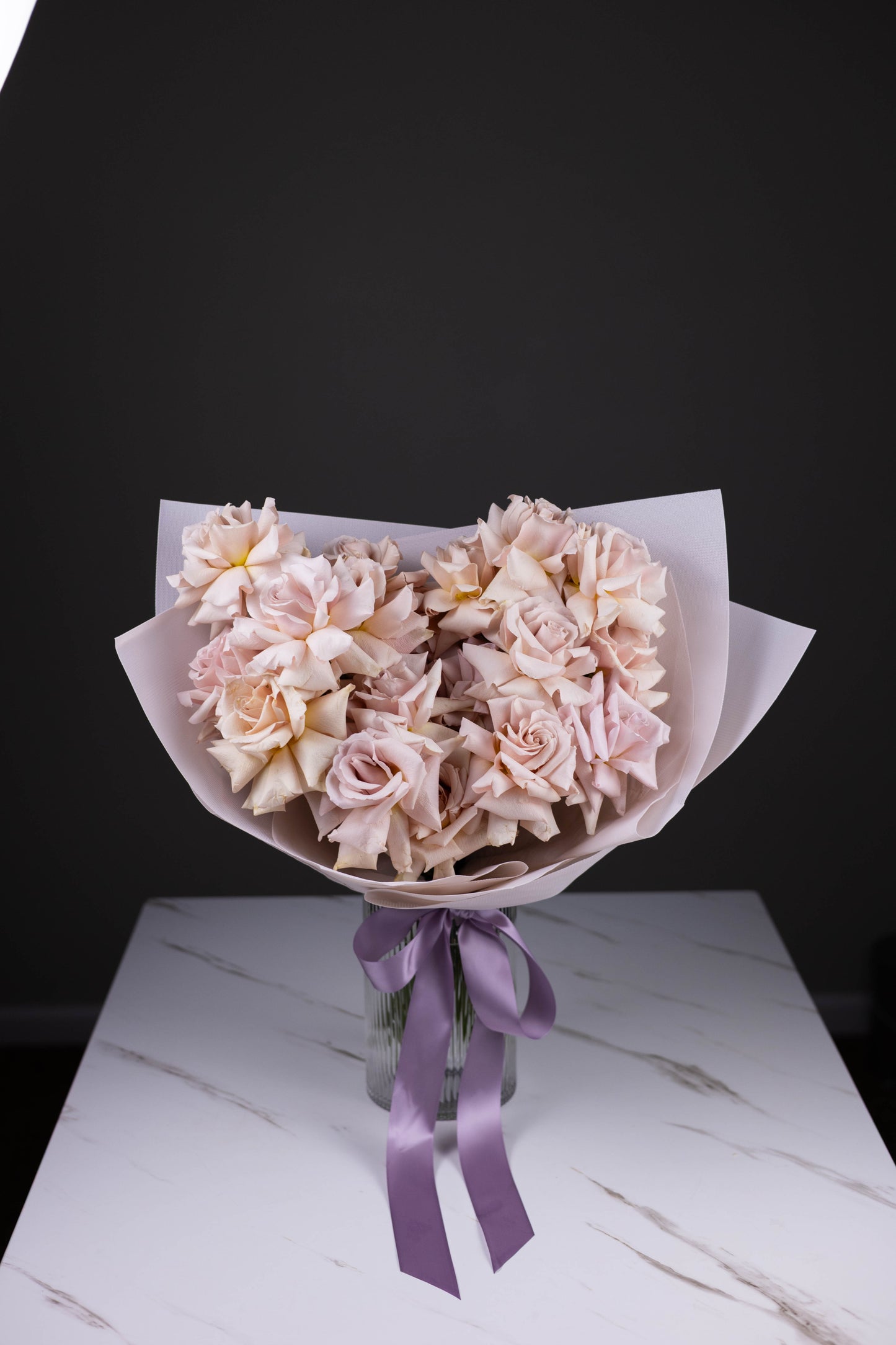 A hand-tied bouquet of blush-lavender roses in pastel wrap, displayed in a fluted glass vase with a lavender satin ribbon