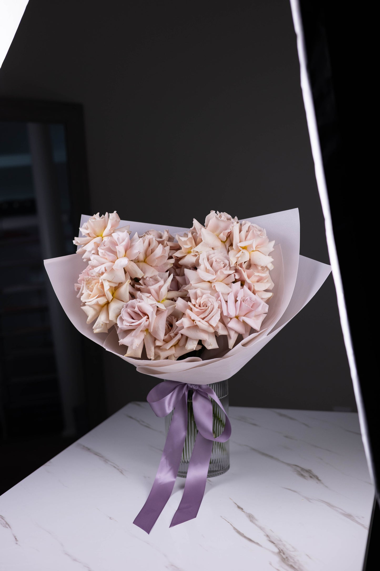 Full bouquet of lavender-toned roses viewed from the front, wrapped in soft pink paper and placed in a vase