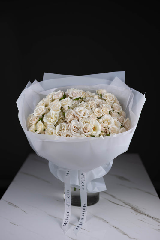 Front view of a voluminous bouquet of pale white roses, tightly packed in translucent paper wrap, resting on a marble surface with signature Maison d’Fleur ribbons.
