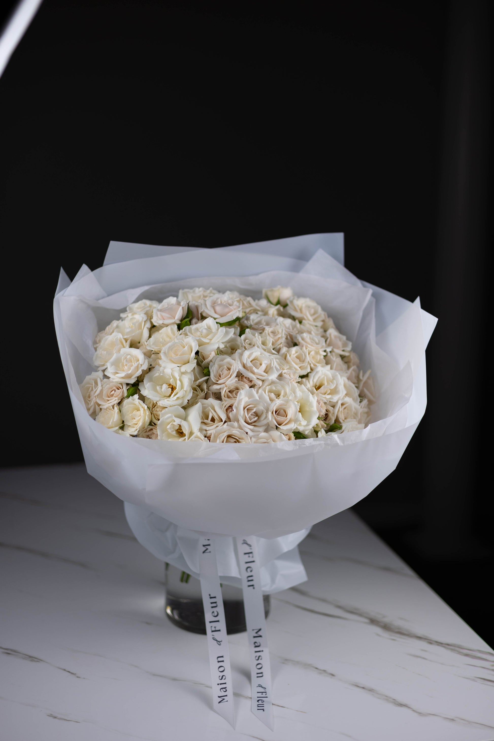 Side angle view of a full round bouquet of soft white and blush roses in sheer white wrapping, tied with Maison d’Fleur branded ribbons.