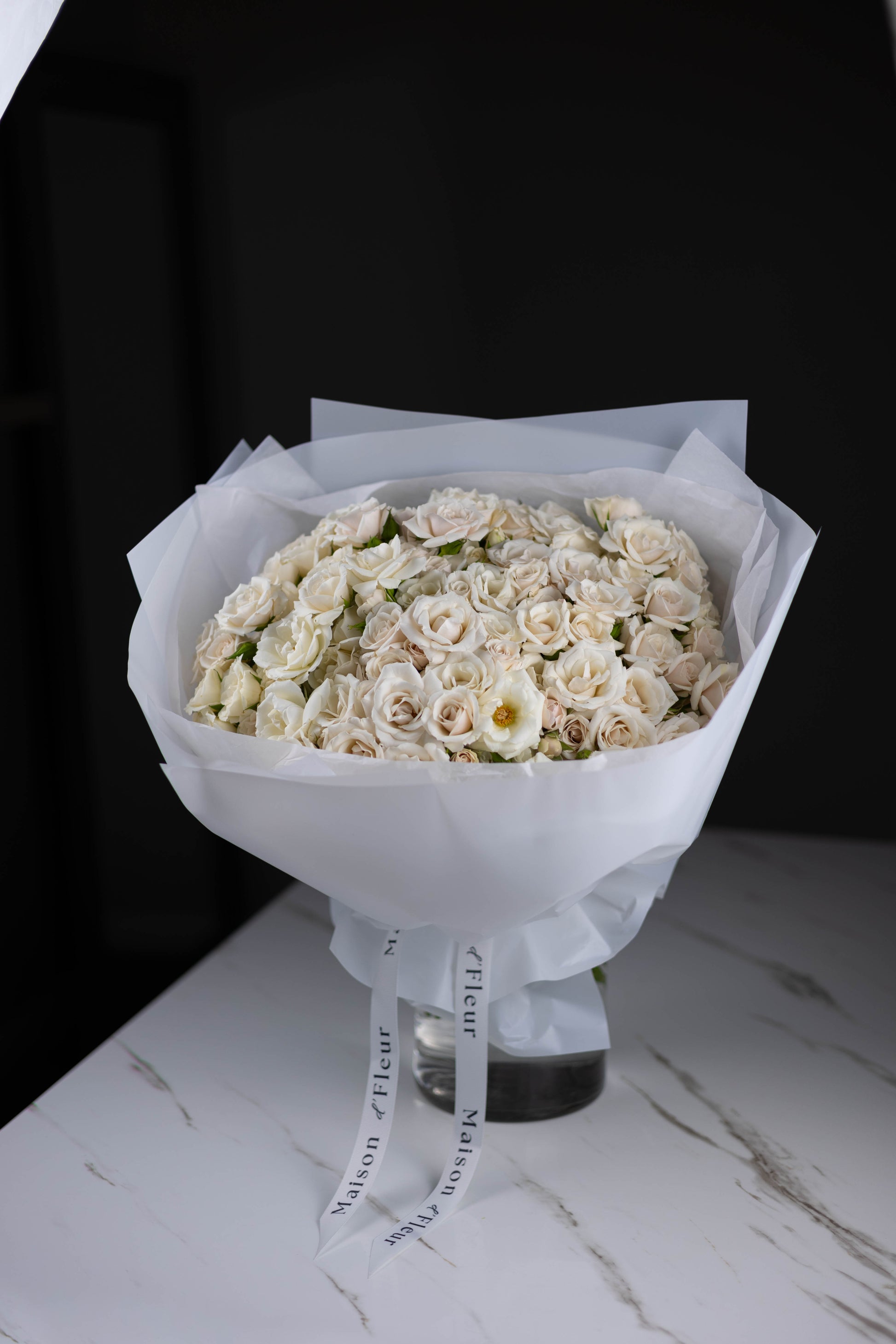 Elegant white rose bouquet wrapped in white paper, displayed in a clear glass vase on a marble table with Maison d’Fleur ribbons.