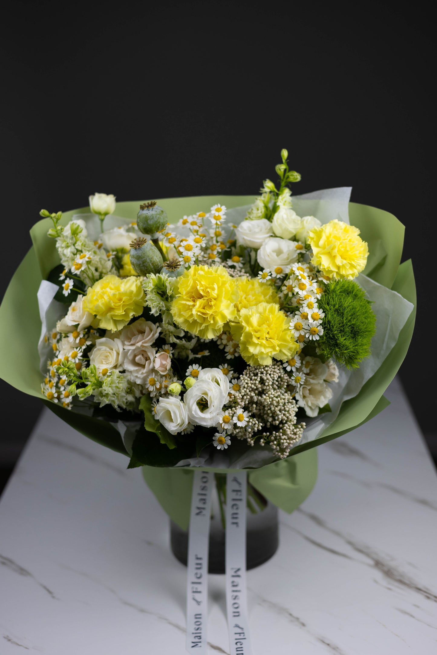 Charming arrangement of yellow and white flowers — carnations, chamomile, and roses — with visible structure and texture, wrapped in matte green floral paper with branded ribbons.
