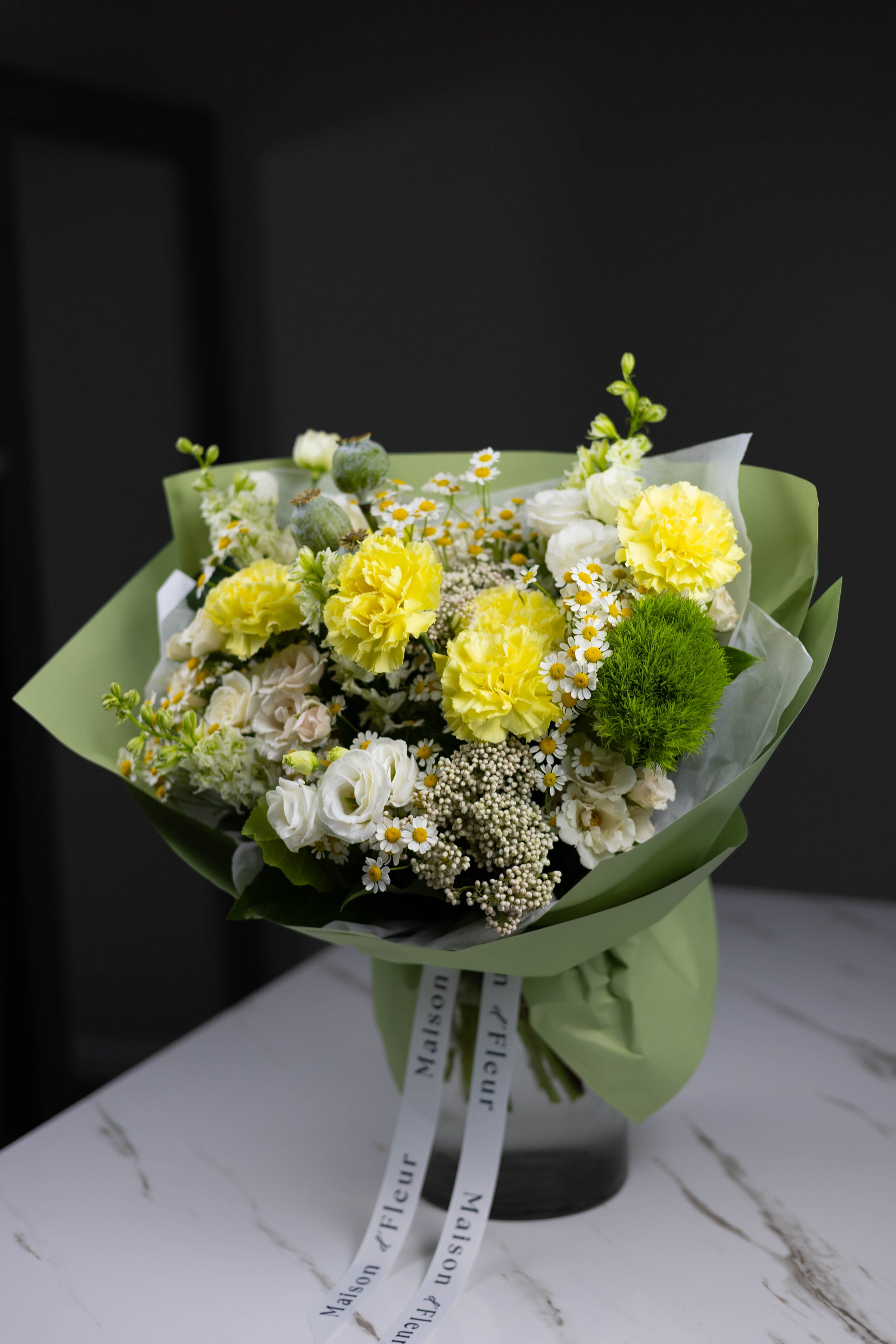 Side view of a lush flower bouquet combining sunny yellow carnations, white florals, soft greenery, and delicate chamomile, all wrapped in pastel green paper.