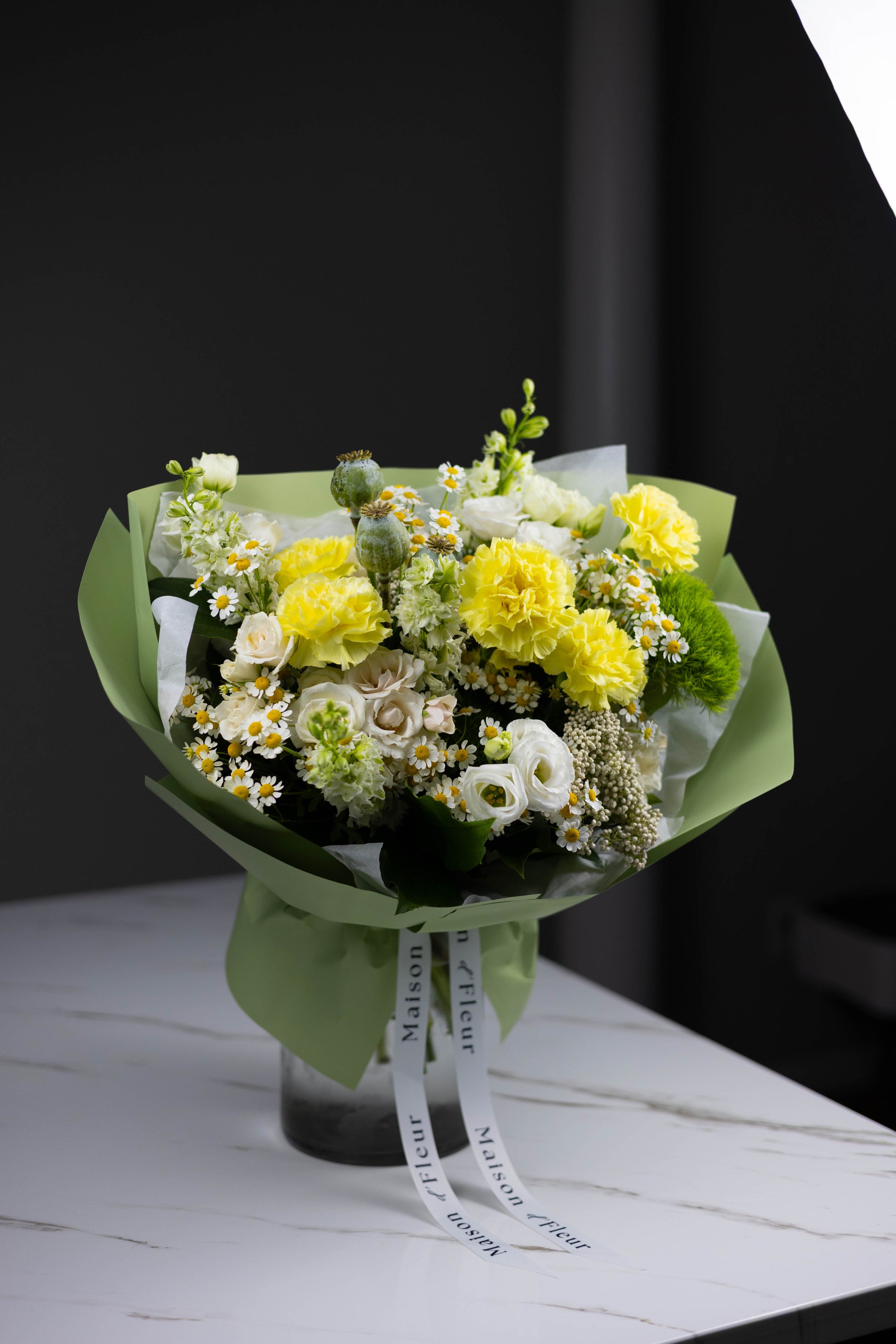 Full-styled bouquet displayed in a clear glass vase, featuring yellow carnations, daisies, white roses, lisianthus, and Maison d’Fleur signature ribbons — presented on a white marble surface with studio lighting.