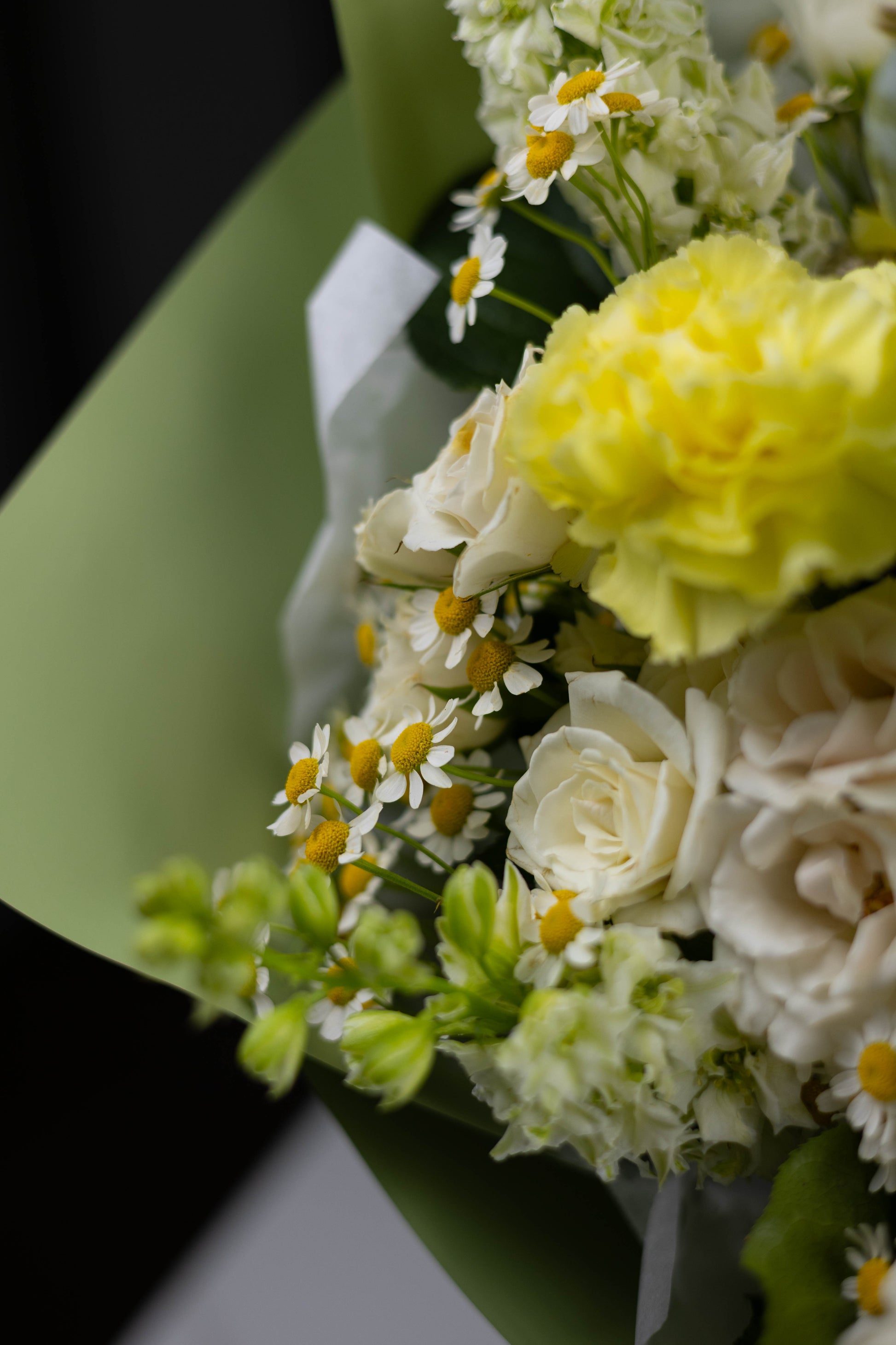 Softly lit bouquet with yellow carnations, chamomile daisies, white roses, and lisianthus, arranged in light green wrapping paper — a cheerful and natural design evoking early summer fields.