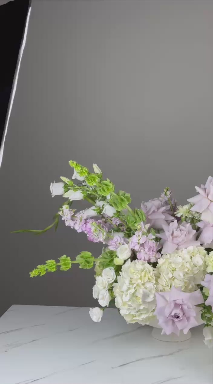 A wide-angle shot of a pastel garden arrangement featuring lavender roses, ivory hydrangeas, and soft greenery in a white compote vase.