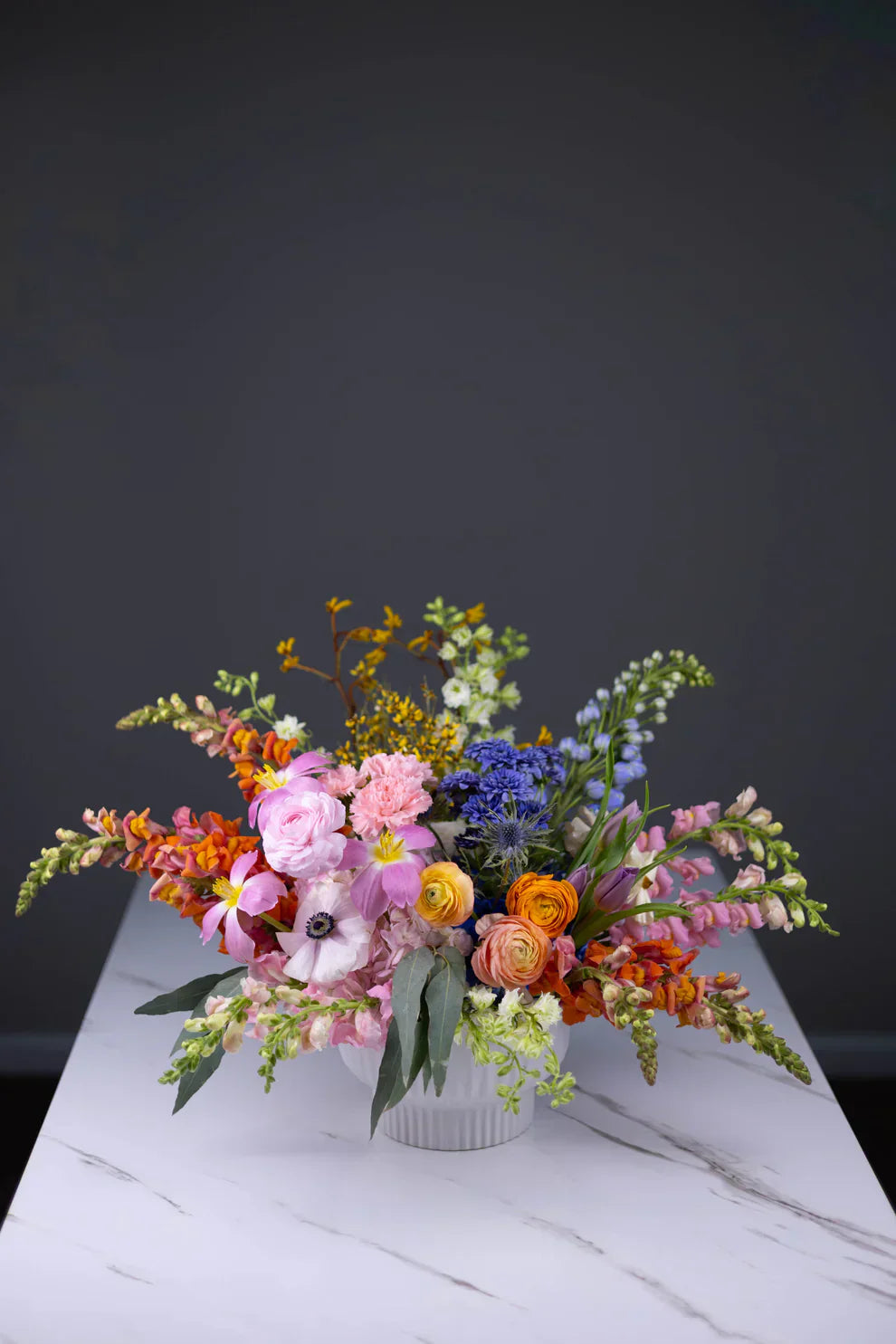 A vibrant floral arrangement with a mix of colorful flowers including blue delphiniums, pink anemones, orange ranunculus, and yellow snapdragons, displayed on a white table with a dark gray background.