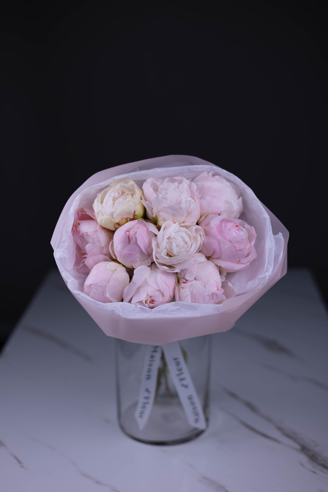 A bouquet of soft blush pink peonies wrapped in white paper, displayed in a clear glass vase on a marble surface, set against a black background.