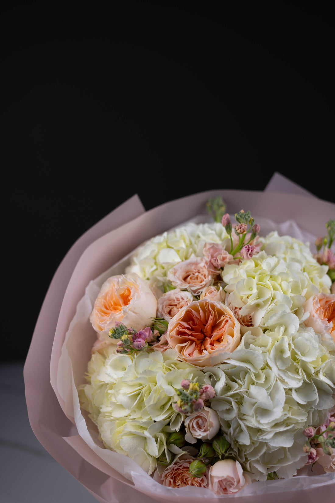 A romantic bouquet of white hydrangeas, peach garden roses, and delicate spray roses, accented with pink buds and soft wrapping, captured in soft lighting.