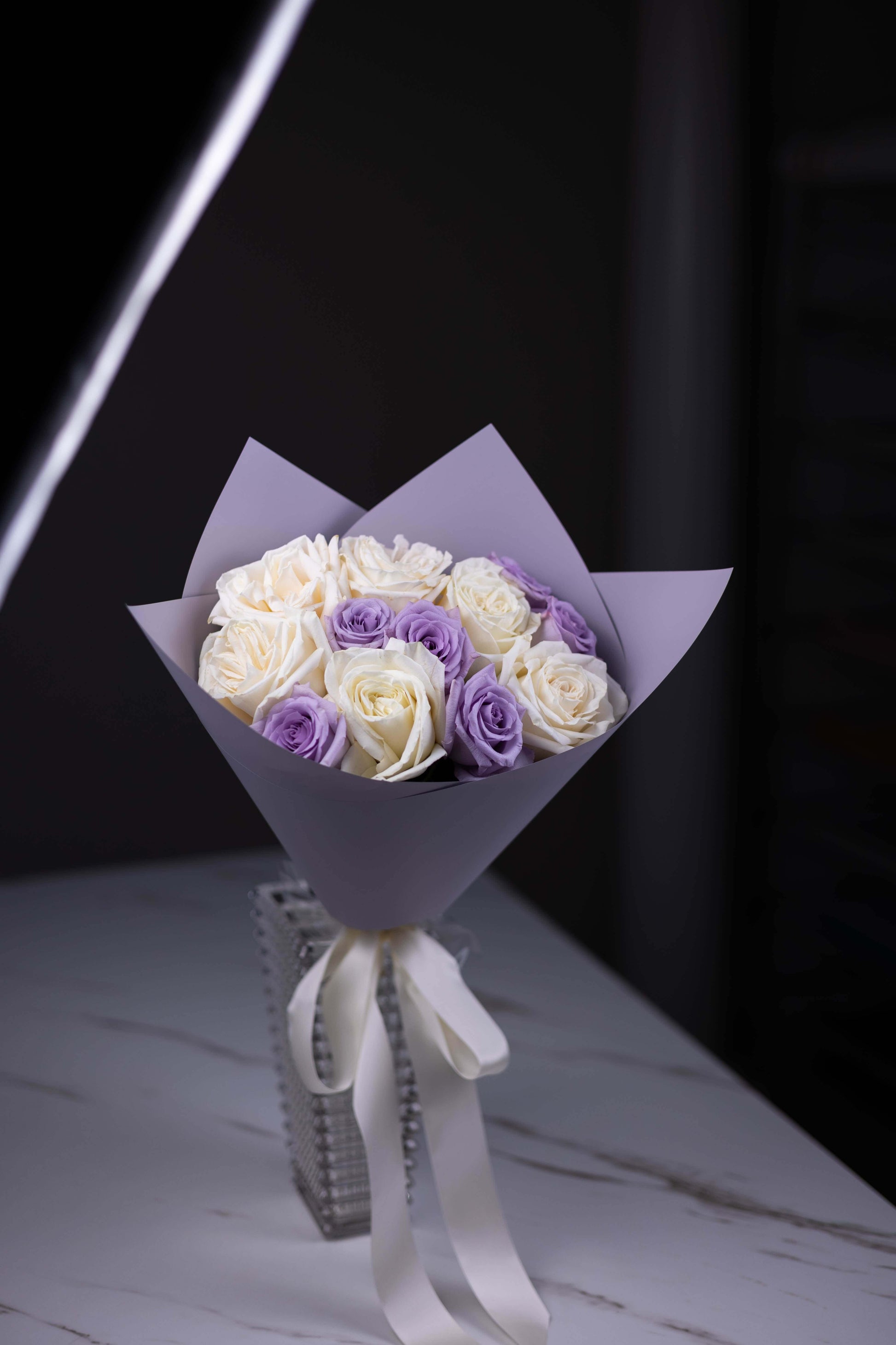 Bouquet of white and purple roses wrapped in purple paper with a white ribbon on a dark background