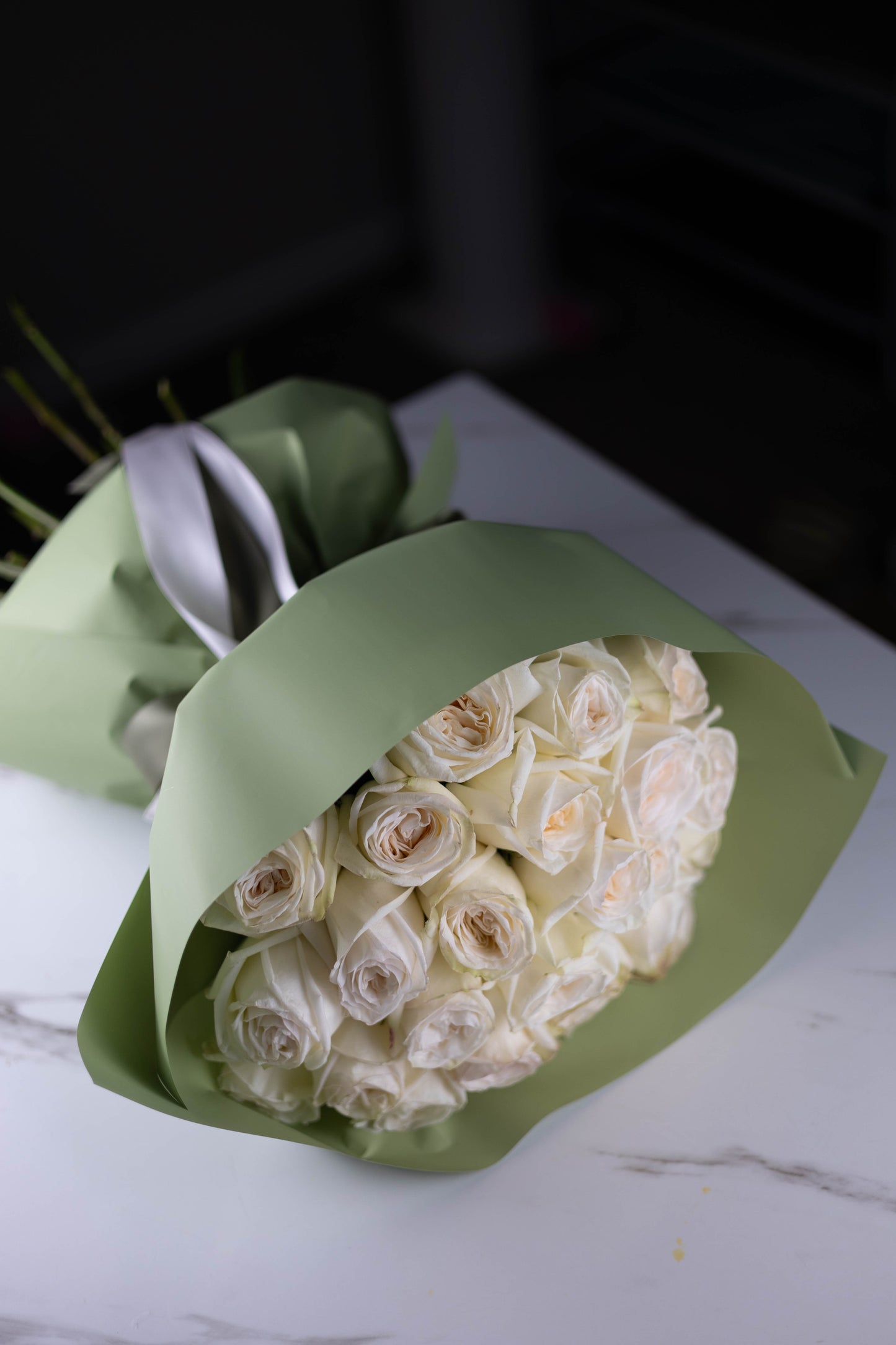 White rose bouquet wrapped in sage-green paper, placed on a white marble surface, showcasing elegant satin ribbon ties.