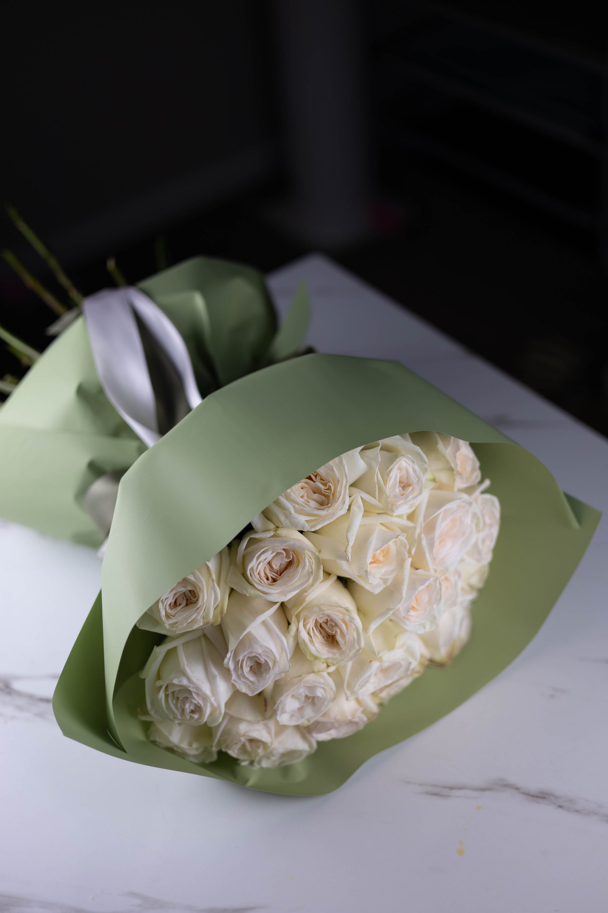 White rose bouquet wrapped in sage-green paper, placed on a white marble surface, showcasing elegant satin ribbon ties.