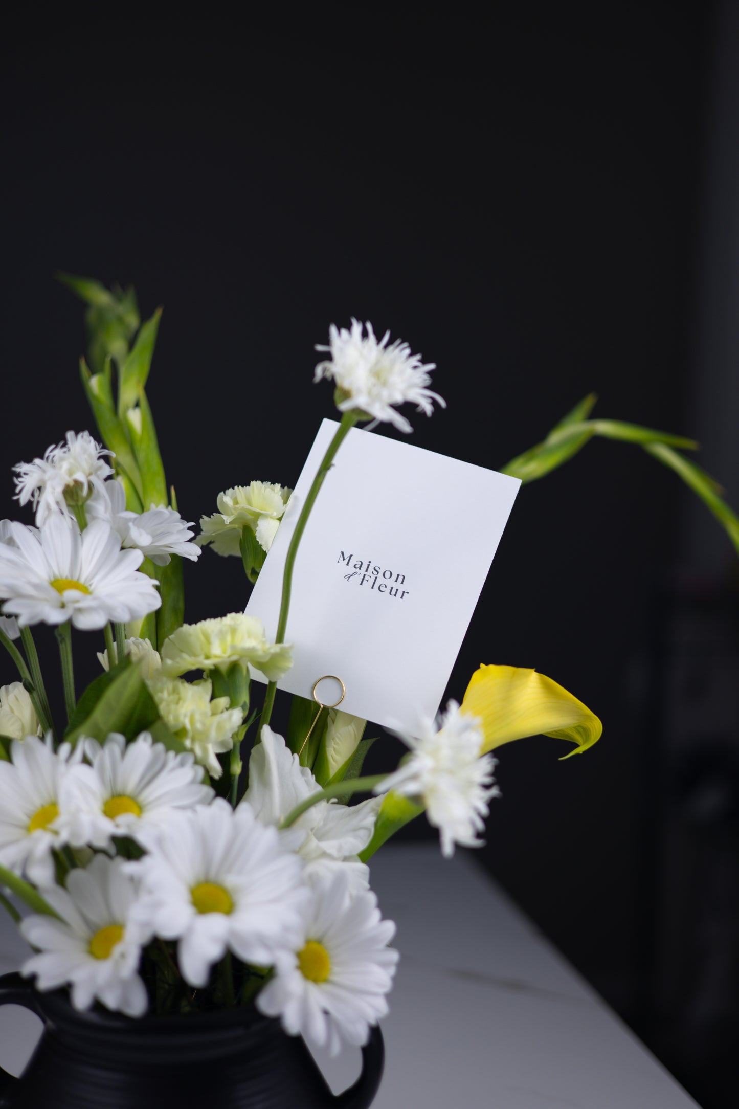 Elegant floral arrangement with white daisies, yellow calla lilies, and green carnations, Maison d’Fleur card visible in the bouquet.