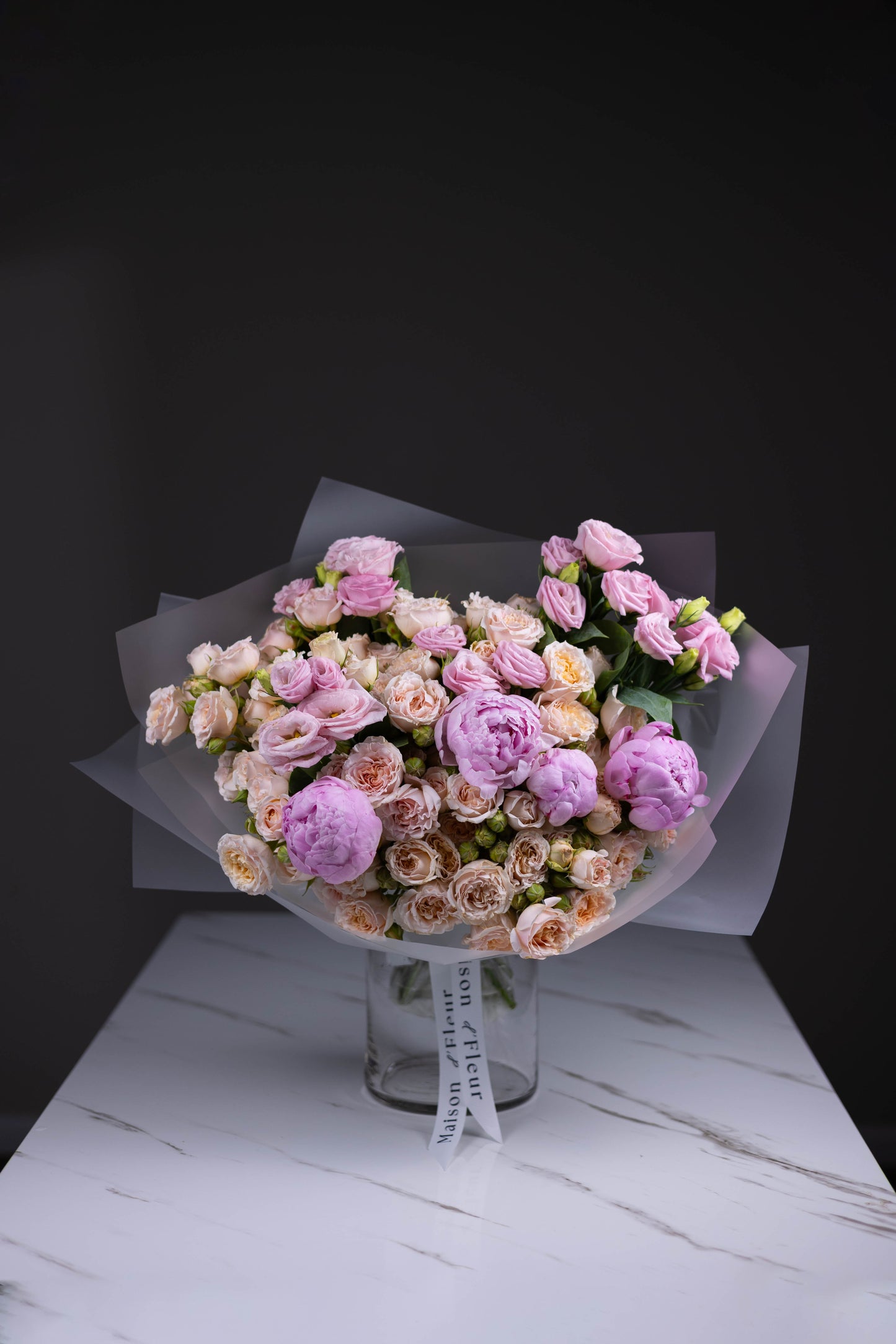Delicate bouquet of blush spray roses, lavender lisianthus, and soft pink peonies wrapped in translucent paper, standing in a Maison d’Fleur branded glass vase.