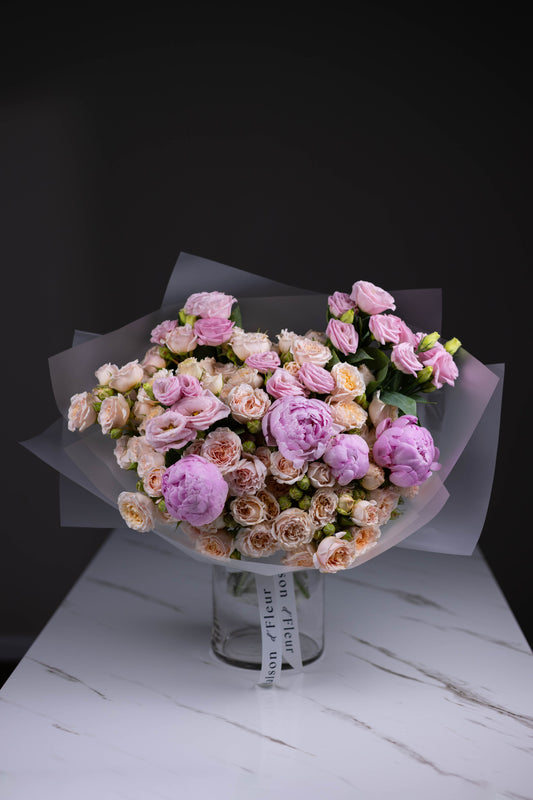 Delicate bouquet of blush spray roses, lavender lisianthus, and soft pink peonies wrapped in translucent paper, standing in a Maison d’Fleur branded glass vase.

